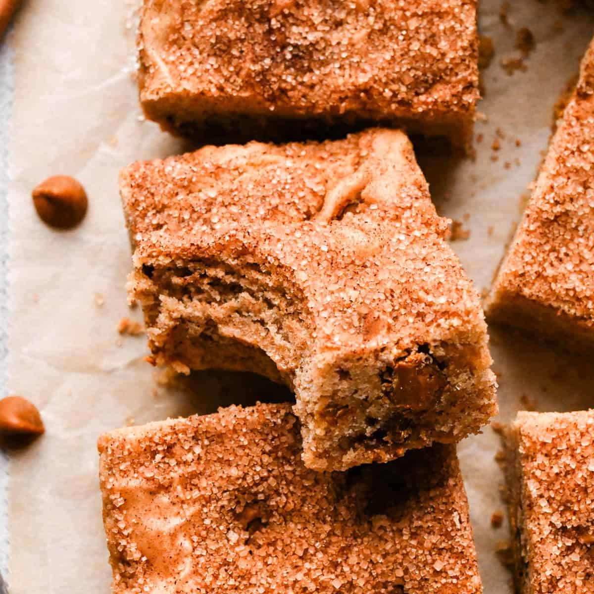 5 snickerdoodle blondies, one resting slightly on top of another one with a bite taken out of it so the chewy texture is visible on parchment paper surrounded by cinnamon chips
