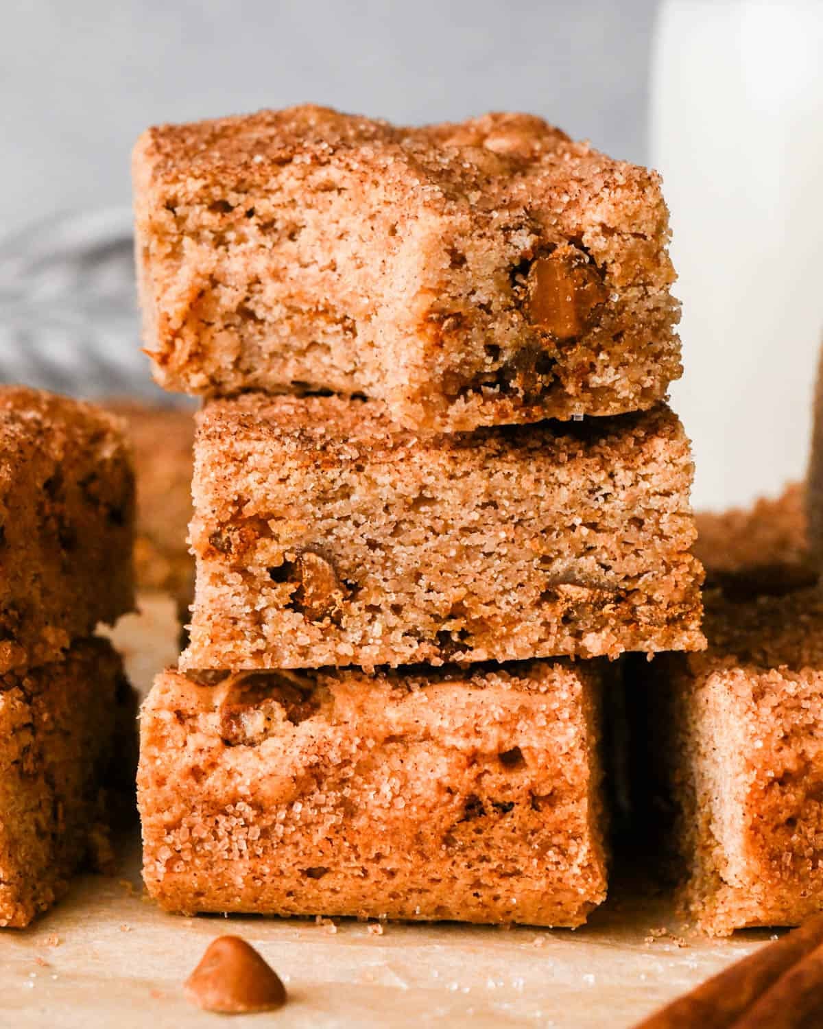 a stack of 3 snickerdoodle blondies, the top one has a bite taken out of it - the chewy texture and cinnamon chips inside are visible. The stack is on parchment paper and surrounded by more blondies, cinnamon chips and a cinnamon stick