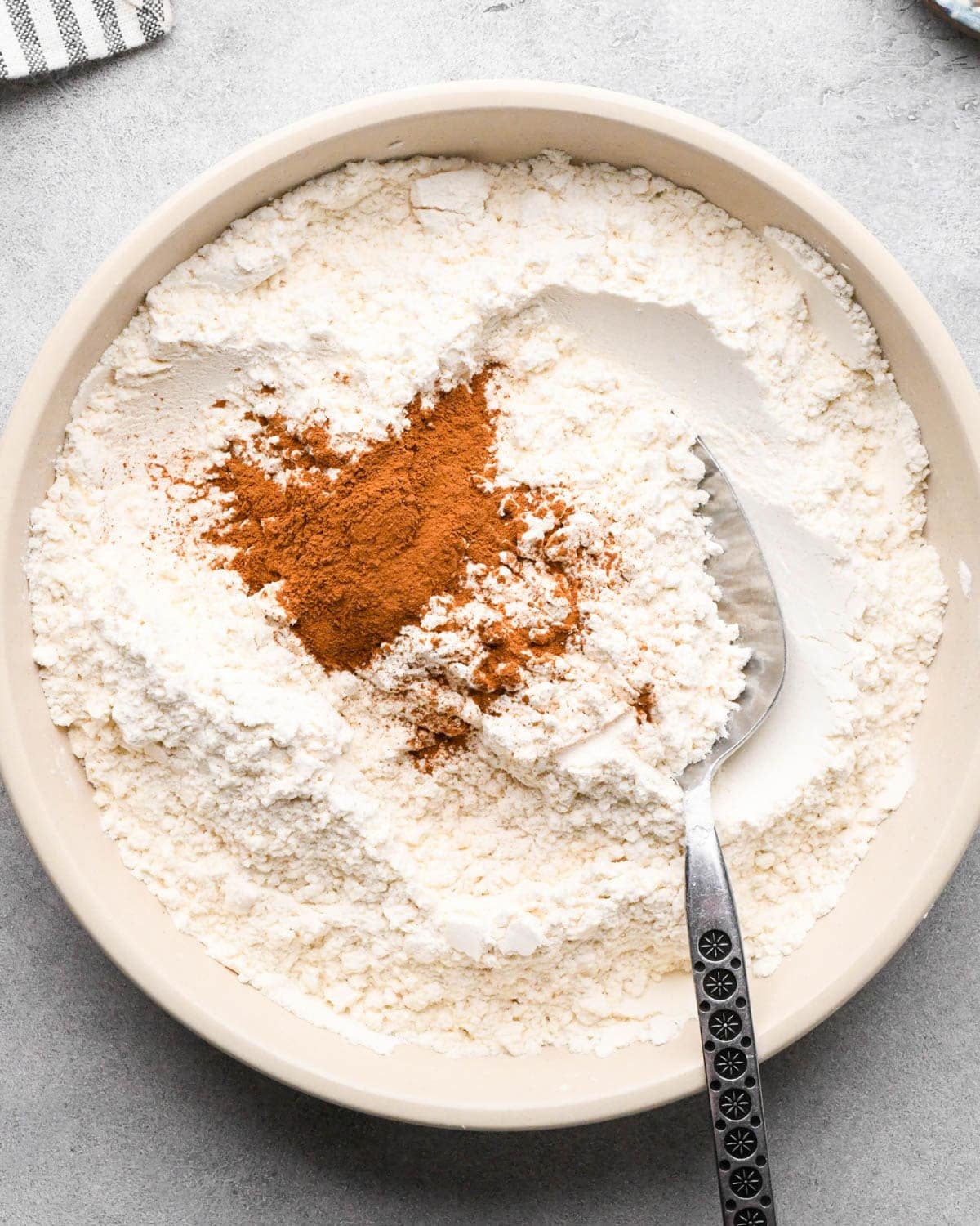 making snickerdoodle blondies -flour, cream of tartar, baking soda, cinnamon and salt in a white bowl with a silver spoon before mixing