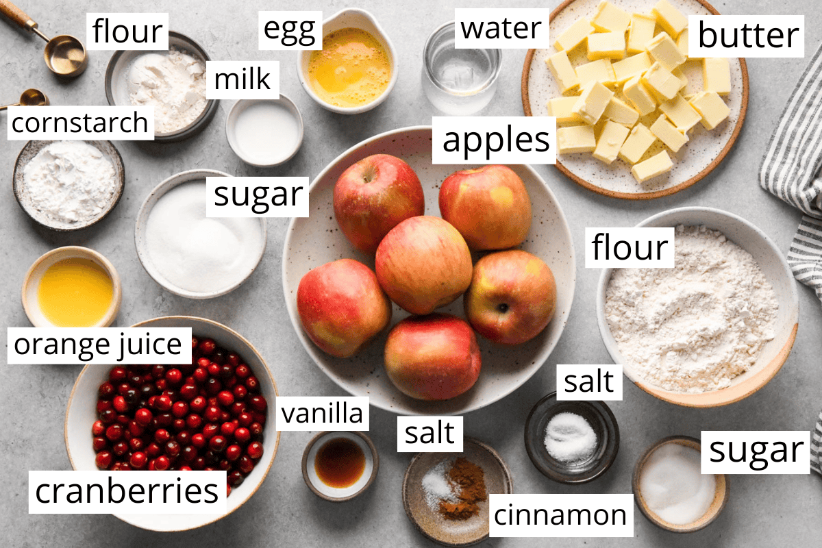 overhead photo of the ingredients in this apple and cranberry pie recipe all in bowls and labeled like apples, butter, cranberries, flour, sugar, orange juice and more