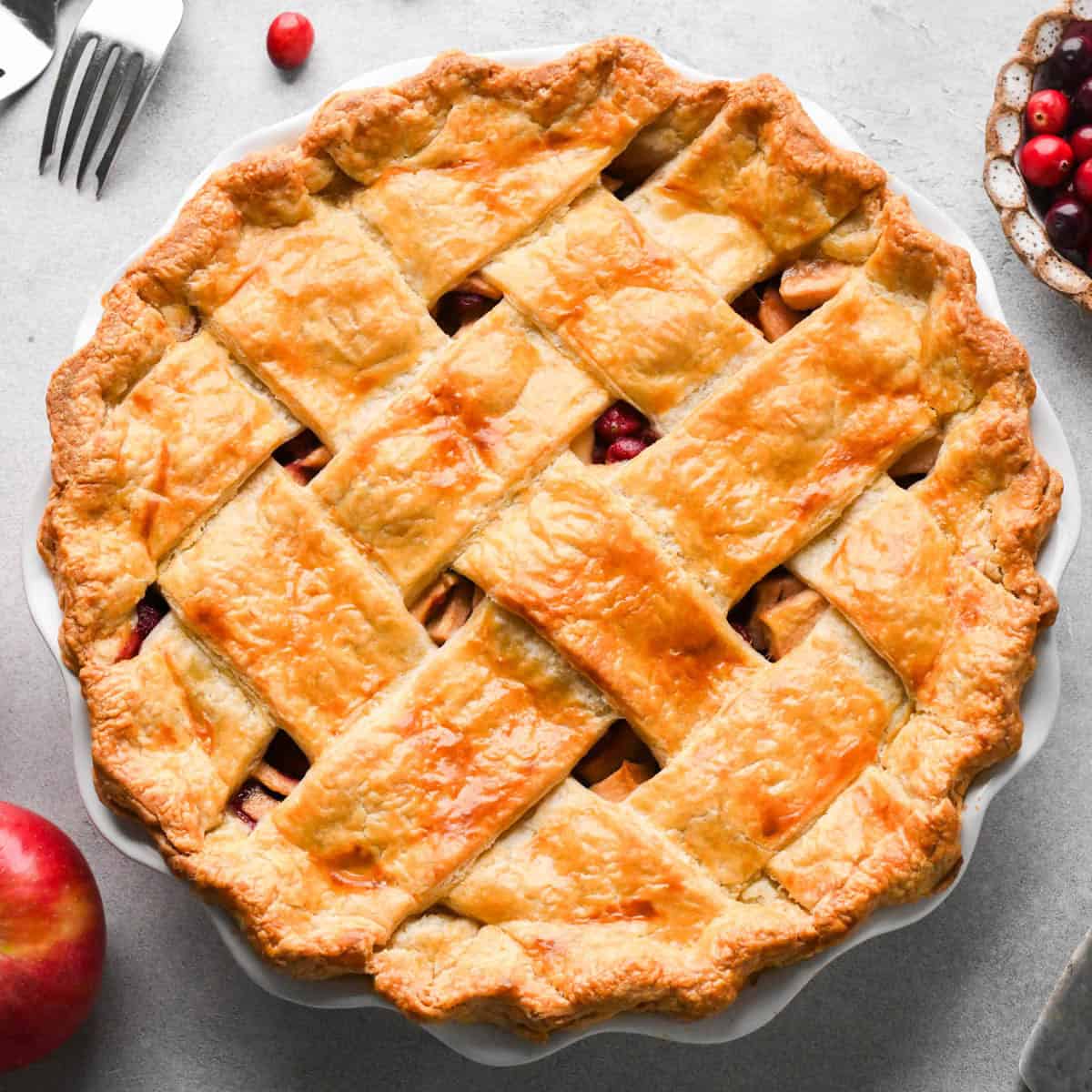 overhead photo of an apple cranberry pie with a lattice crust surrounded by forks, cranberries and an apple