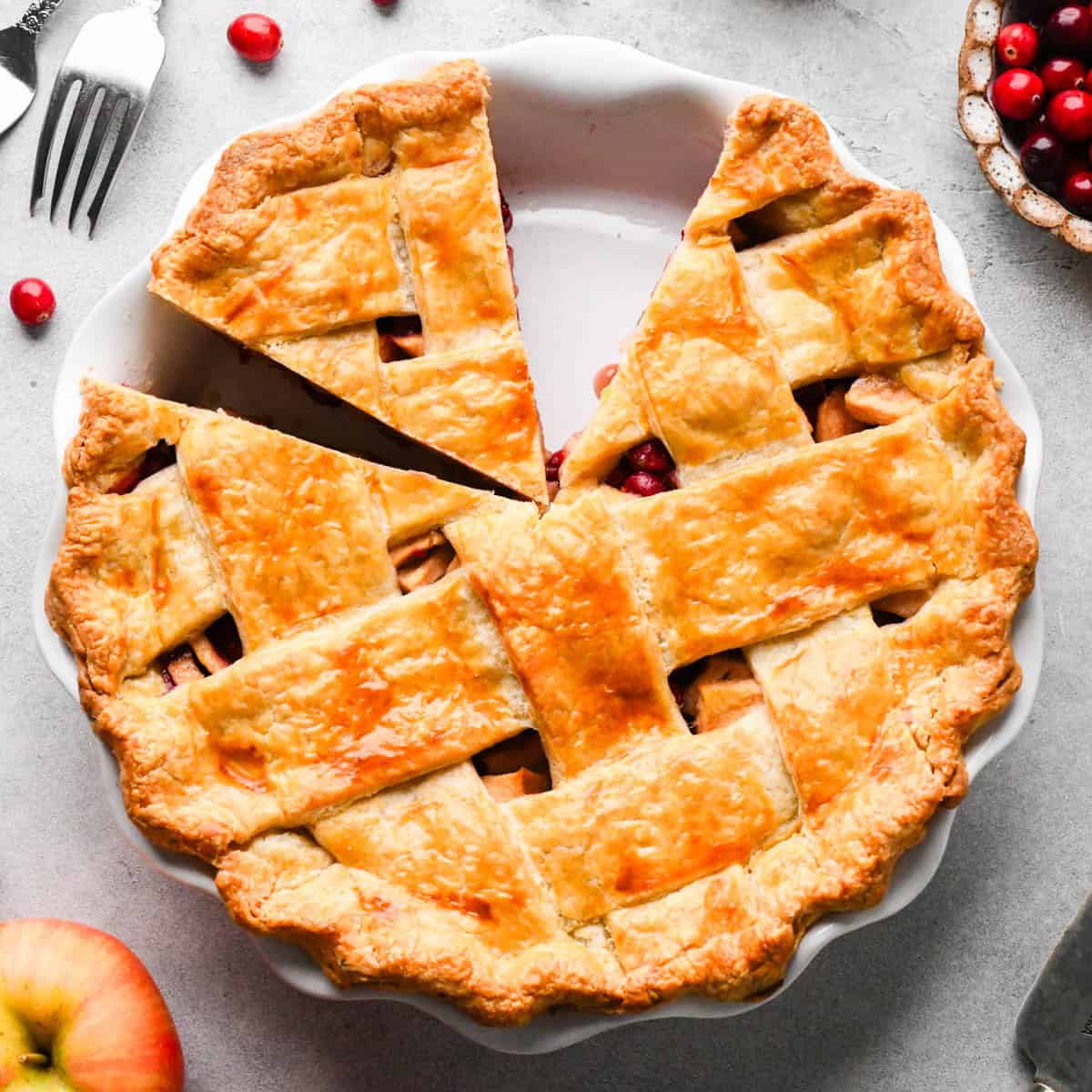 apple cranberry pie with a lattice crust in a white pie dish with a piece cut out of it surrounded by cranberries and an apple and a fork