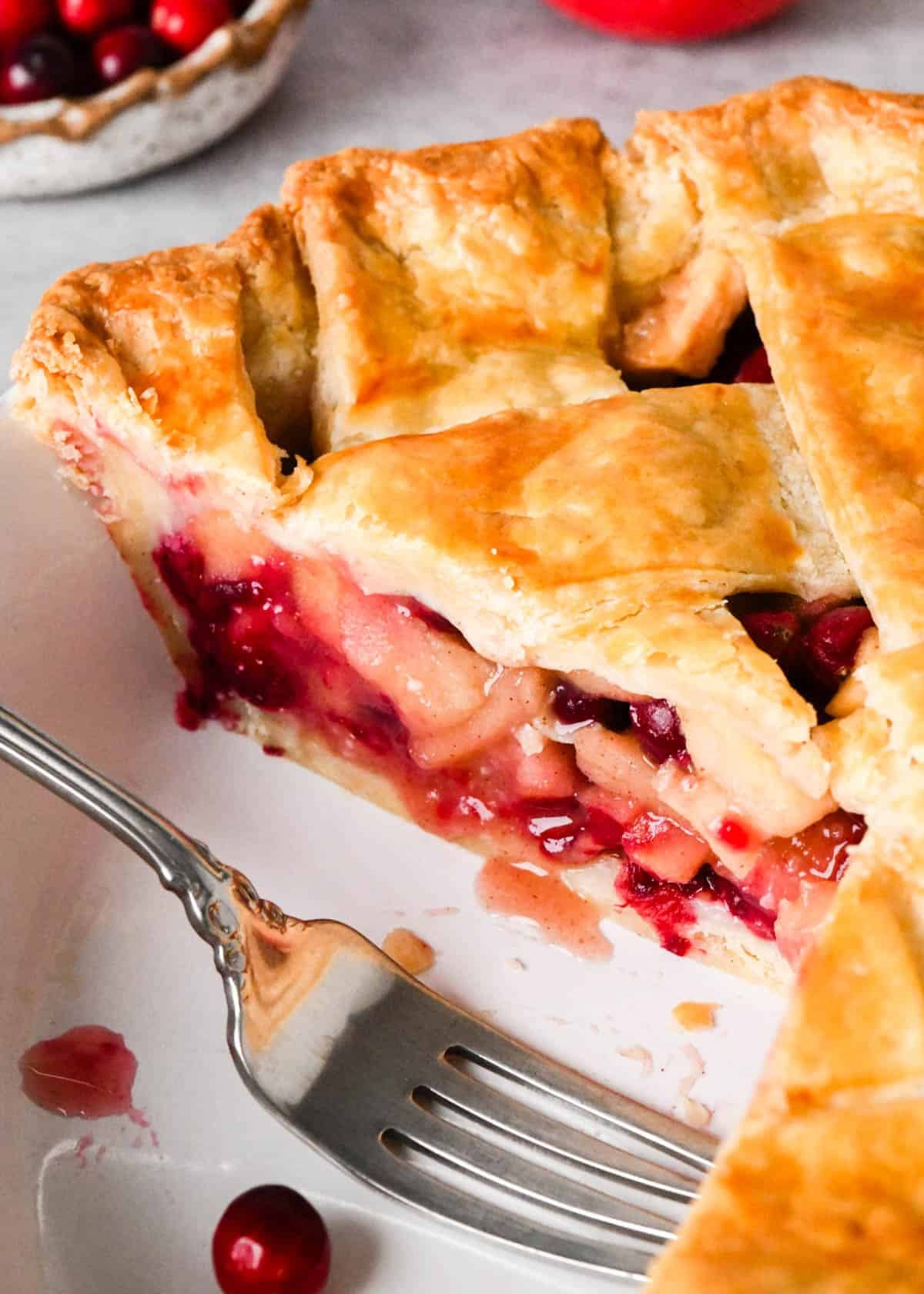 front side view of an apple cranberry pie with a lattice crust in a white pie dish with a fork and a piece cut out of it so you can see the filling