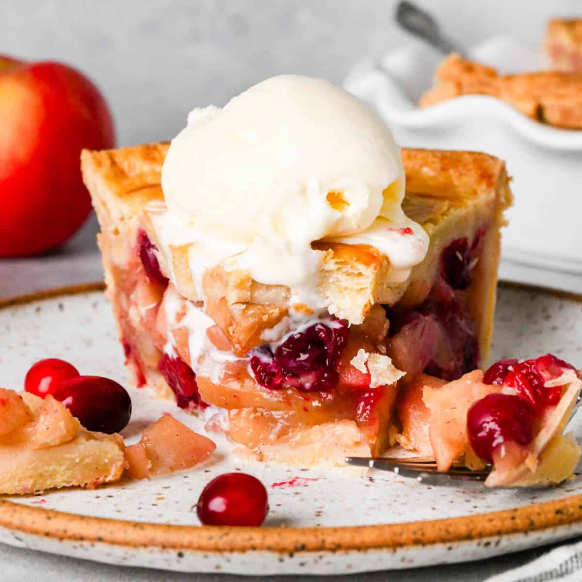 front view a slice of apple cranberry pie on a plate with a scoop of melty vanilla ice cream on top and a bite taken out of it, there are cranberries, apples, a fork and the rest of the pie int he background
