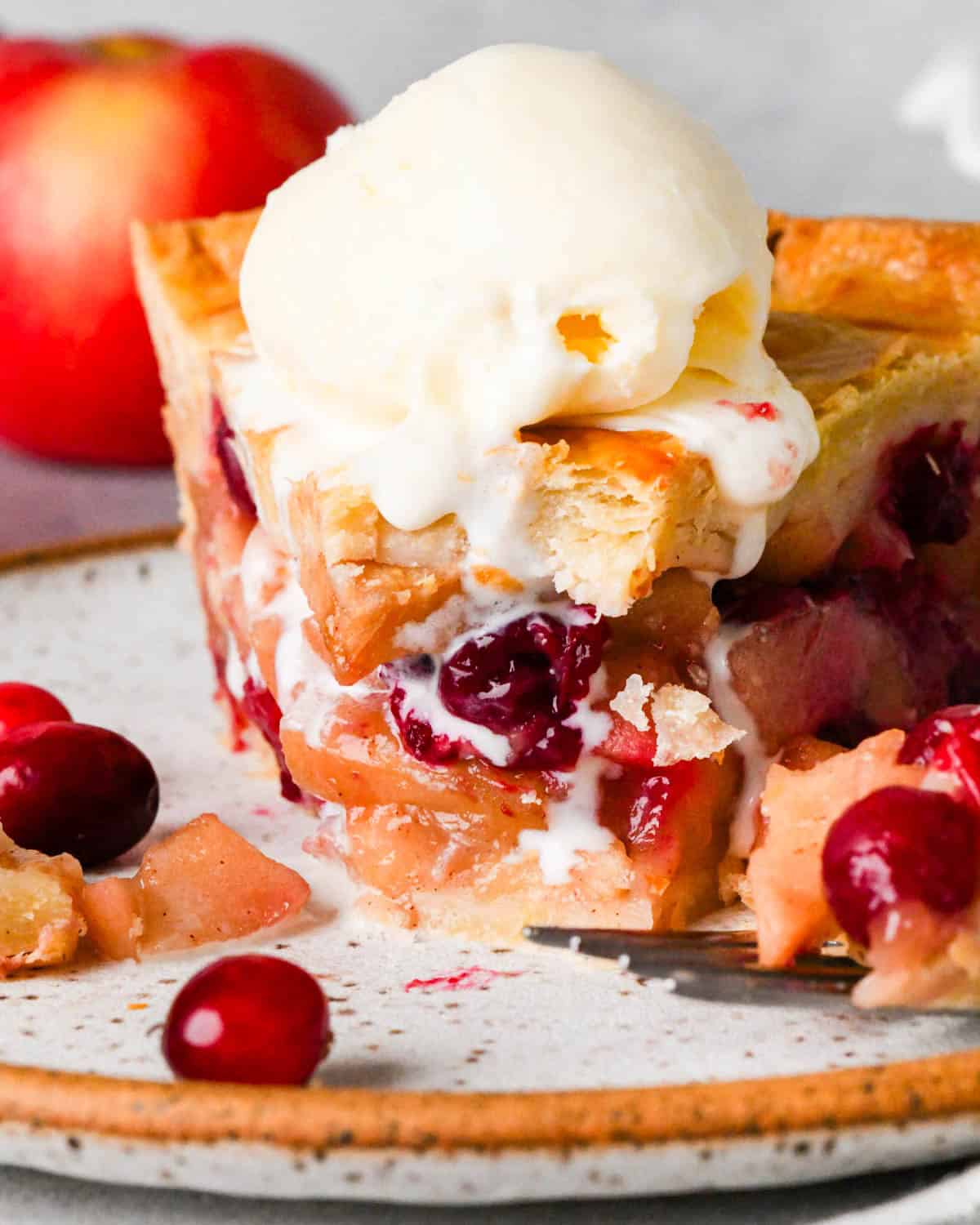 close up front view of a slice of apple cranberry pie on a plate with a taken out of it so the filling is easy to see, with a scoop of melty vanilla ice cream on top, there are cranberries, apples, a fork and the rest of the pie int he background
