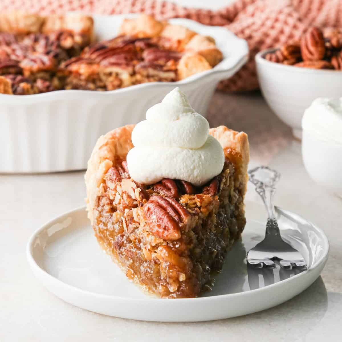 a slice of pecan pie topped with whipped cream on a white plate with a fork so they moist pecan pie filling is visible. The rest of the pecan pie is in a white ceramic pie dish in the background along with a bowl of pecans and a light orange waffle towel.