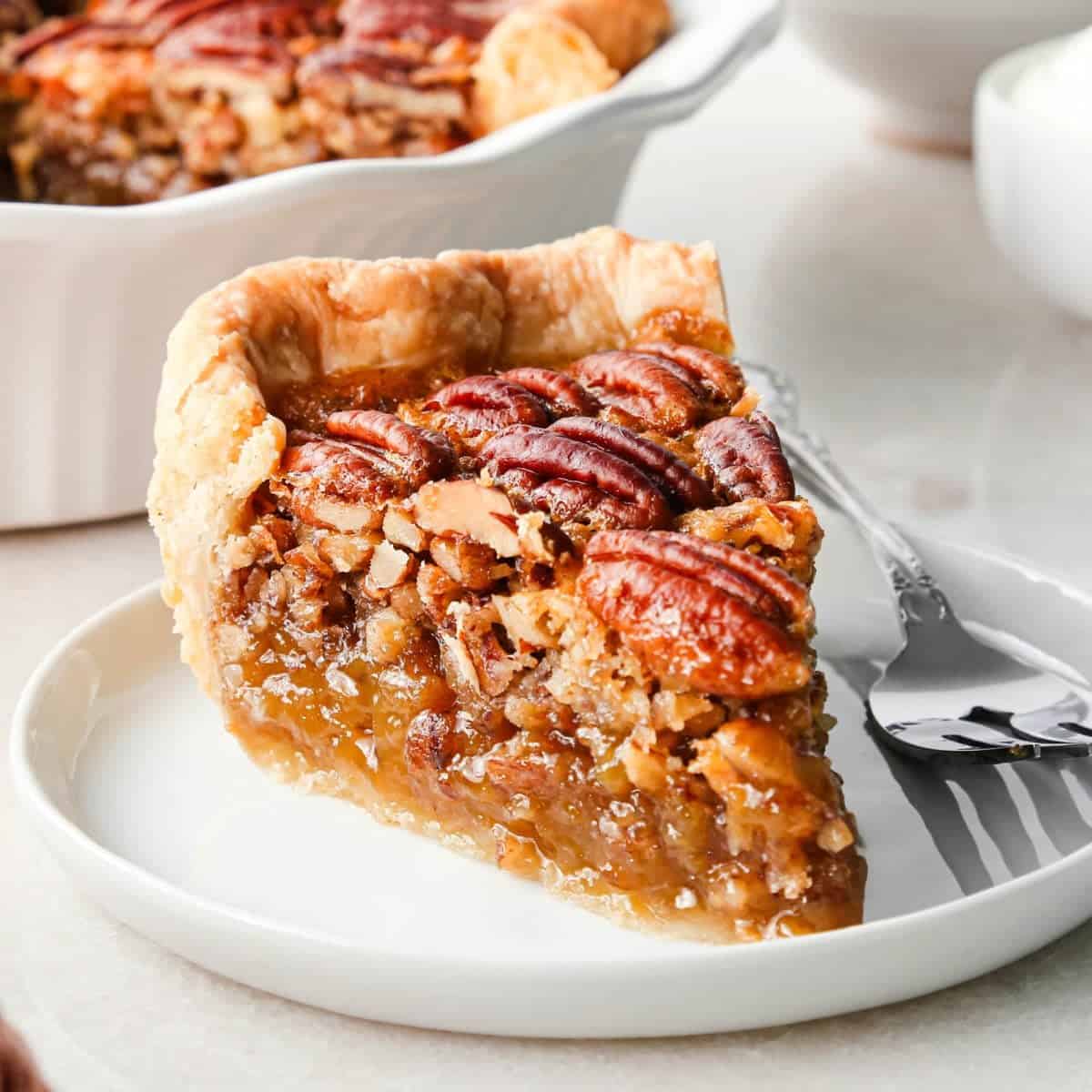 a slice of pecan pie on a white plate with a fork so they moist pecan pie filling is visible. The rest of the pecan pie is in a white ceramic pie dish in the background along with a bowl of whipped cream.