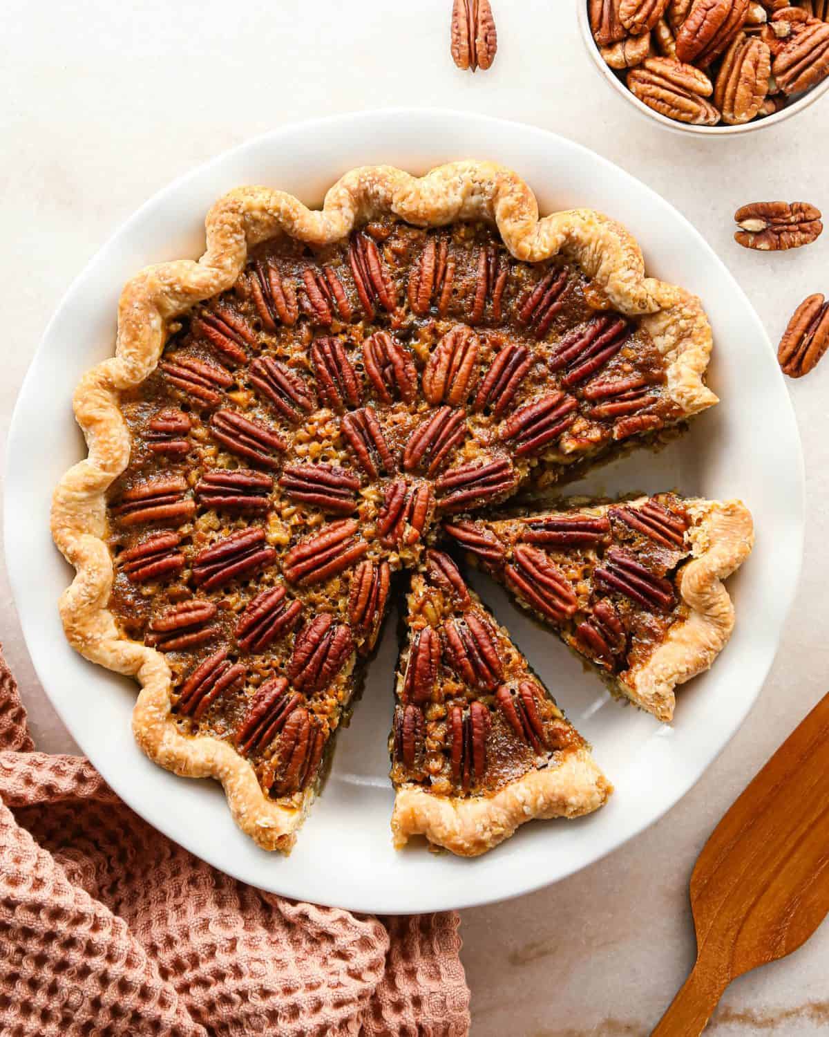 pecan pie in a white ceramic pie dish with a fluted edge crust with two triangular slices cut out of it, there's a light orange waffle towel in the bottom left corner and pecans in the top right corner.
