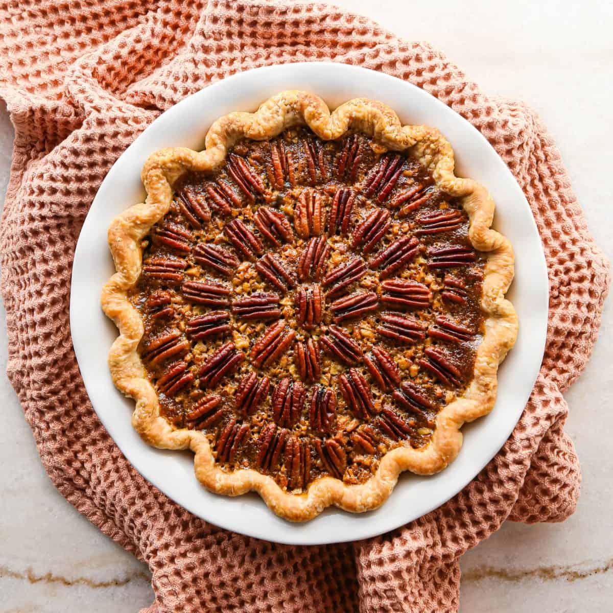 pecan pie in a white ceramic pie dish with a fluted edge crust with two triangular slices cut out of it, there's a light orange waffle towel under the dish
