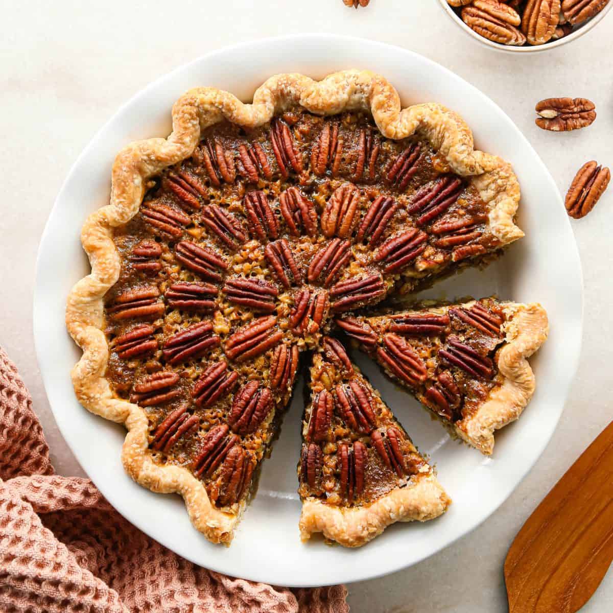 pecan pie in a white ceramic pie dish with a fluted edge crust with two triangular slices cut out of it, there's a light orange waffle towel in the bottom left corner, a wooden pie server in the bottom right corner, and pecans in the top right corner.