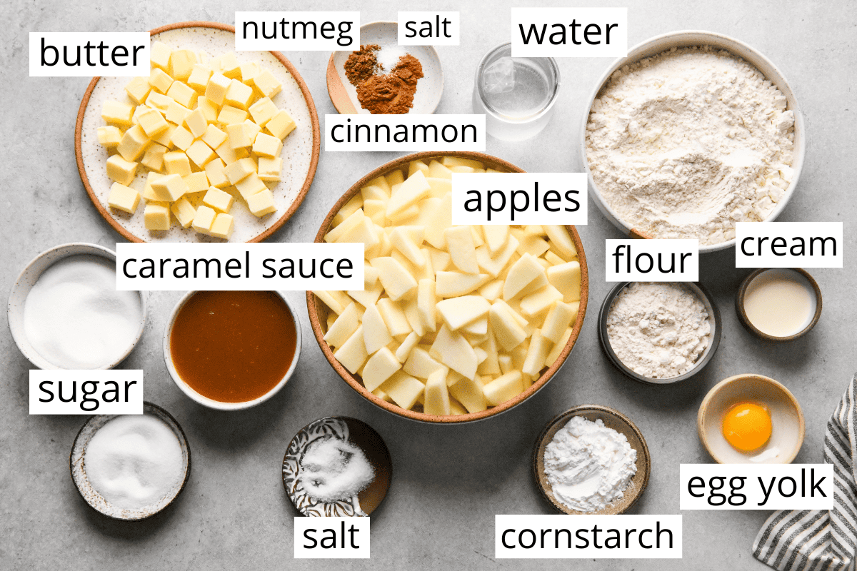 overhead photo of the ingredients in this caramel apple pie recipe all in bowls and labeled like apples, flour, butter, cream, egg, caramel sauce, cinnamon, sugar and more.
