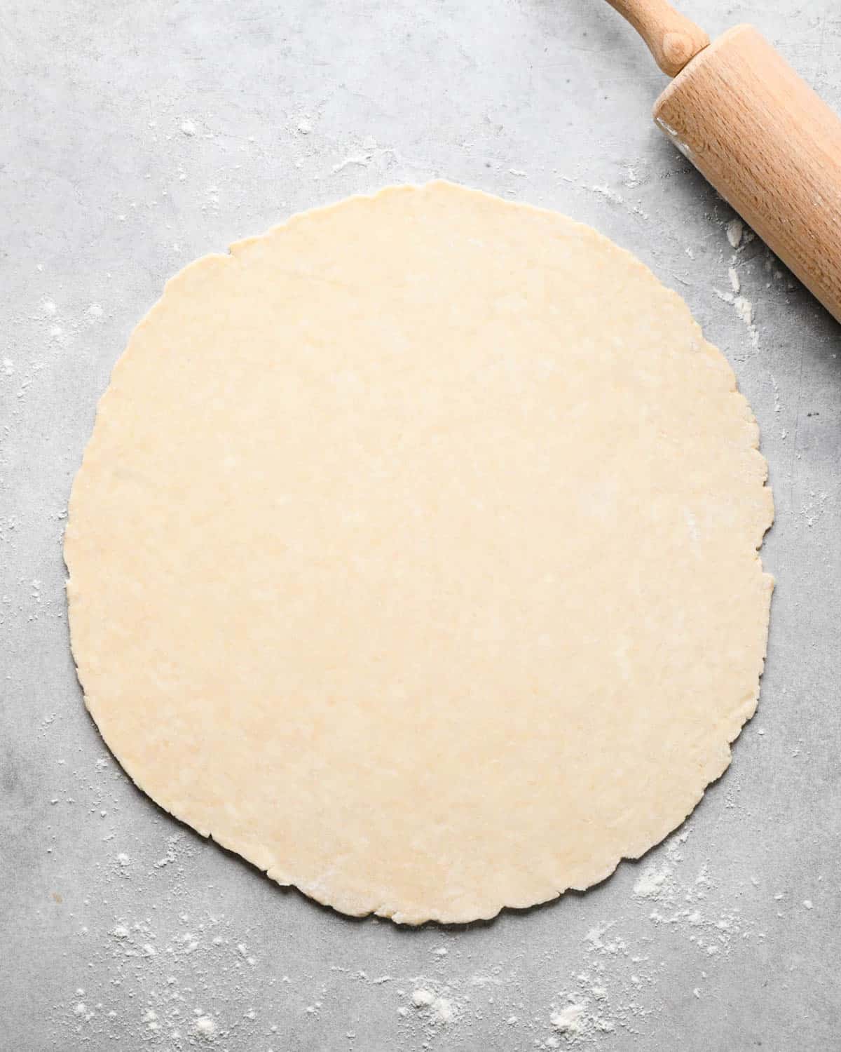 making caramel apple pie - disc of pie dough on a gray, floured surface after rolling out with a wooden rolling pin in the top right corner
