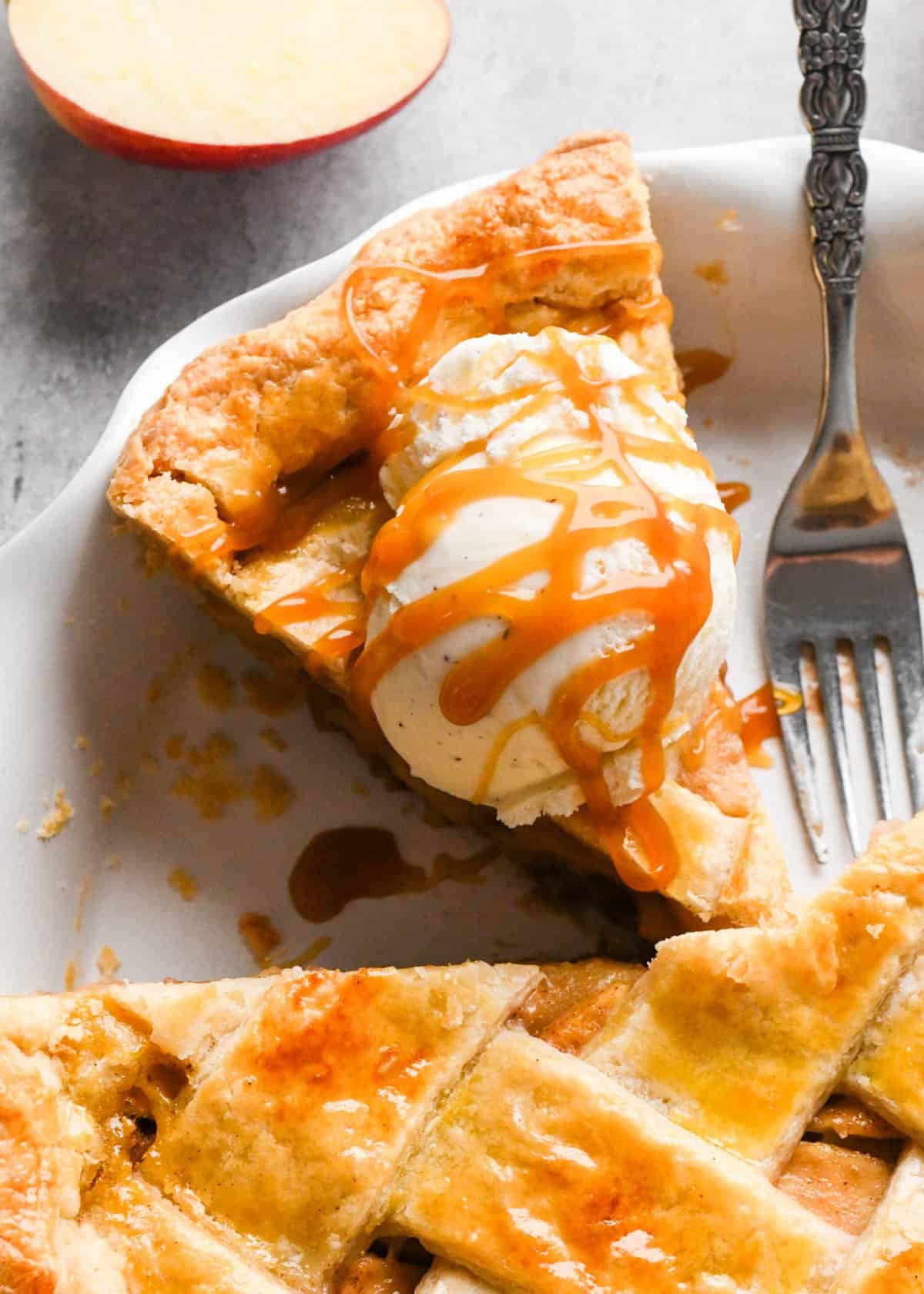 overhead view of a slice of caramel apple pie on in a white pie dish with a coop of vanilla ice cream and caramel sauce drizzled on top of the ice cream. There is a cinnamon fork in the pie dish next to the pie and an apple in the background