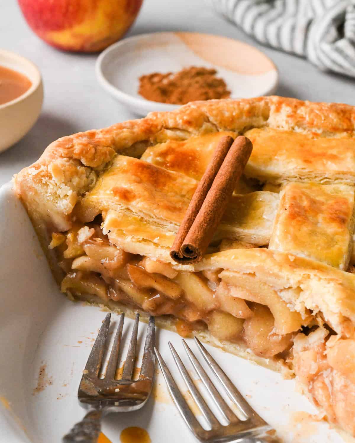 homemade caramel apple pie recipe in a white pie dish with some slices cut out so the thick caramel apple filling is visible. There is a cinnamon stick on top of the pie and two forks in the pie dish with a bowl of caramel sauce, a bowl of cinnamon and an apple in the background