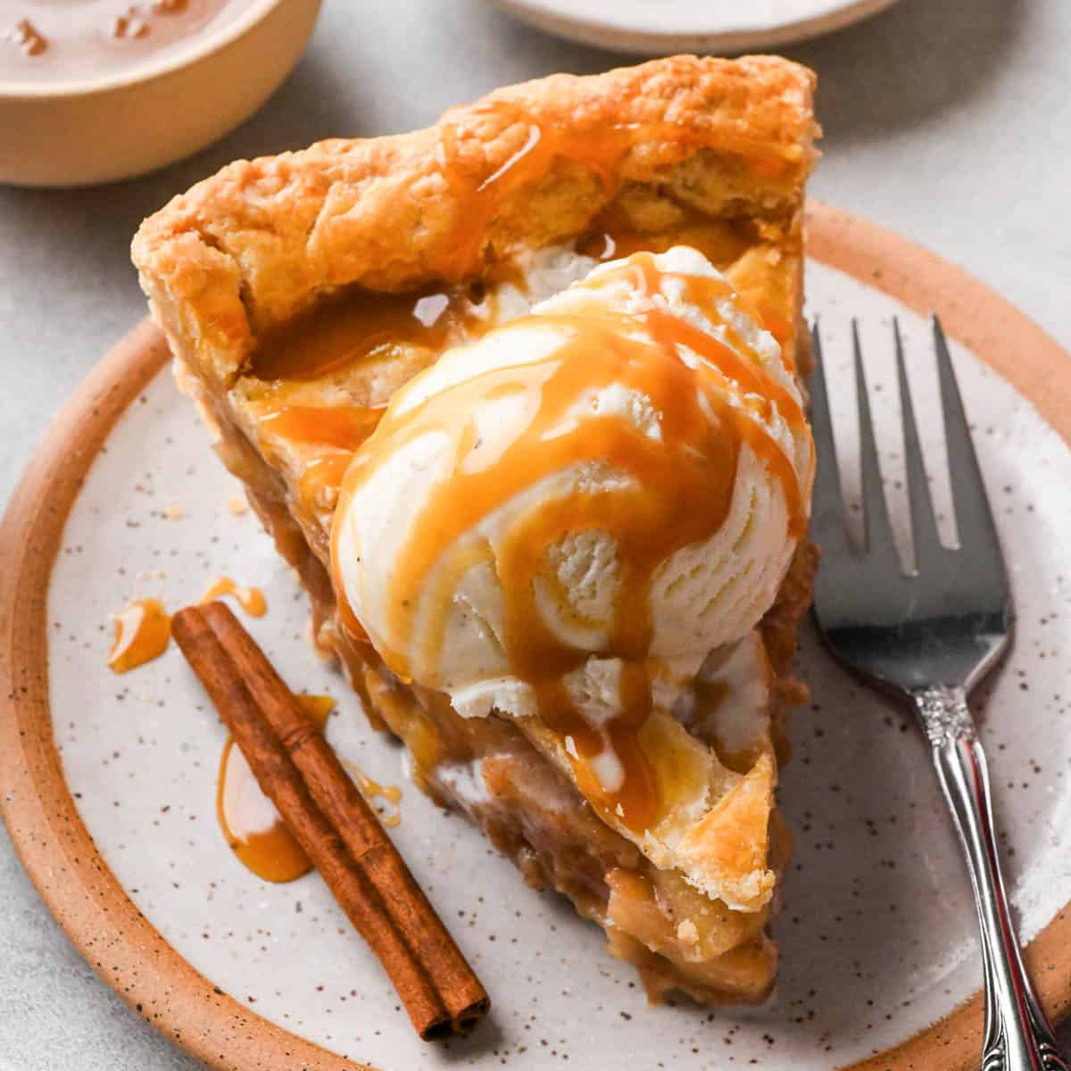 a slice of caramel apple pie on a ceramic plate topped with a scoop of vanilla ice cream and drizzled with caramel sauce. There is a fork and a cinnamon stick on the plate and a bowl of caramel sauce in the background