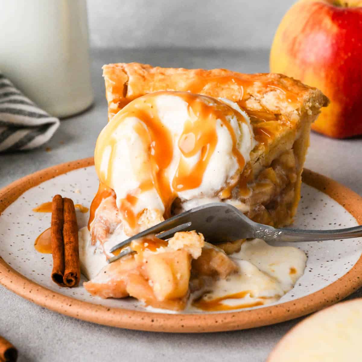a slice of apple pie on a ceramic plate topped with melty vanilla ice cream and caramel sauce with a fork taking a bite out of the pie. There's a cinnamon stick on the plate and an apple in the background
