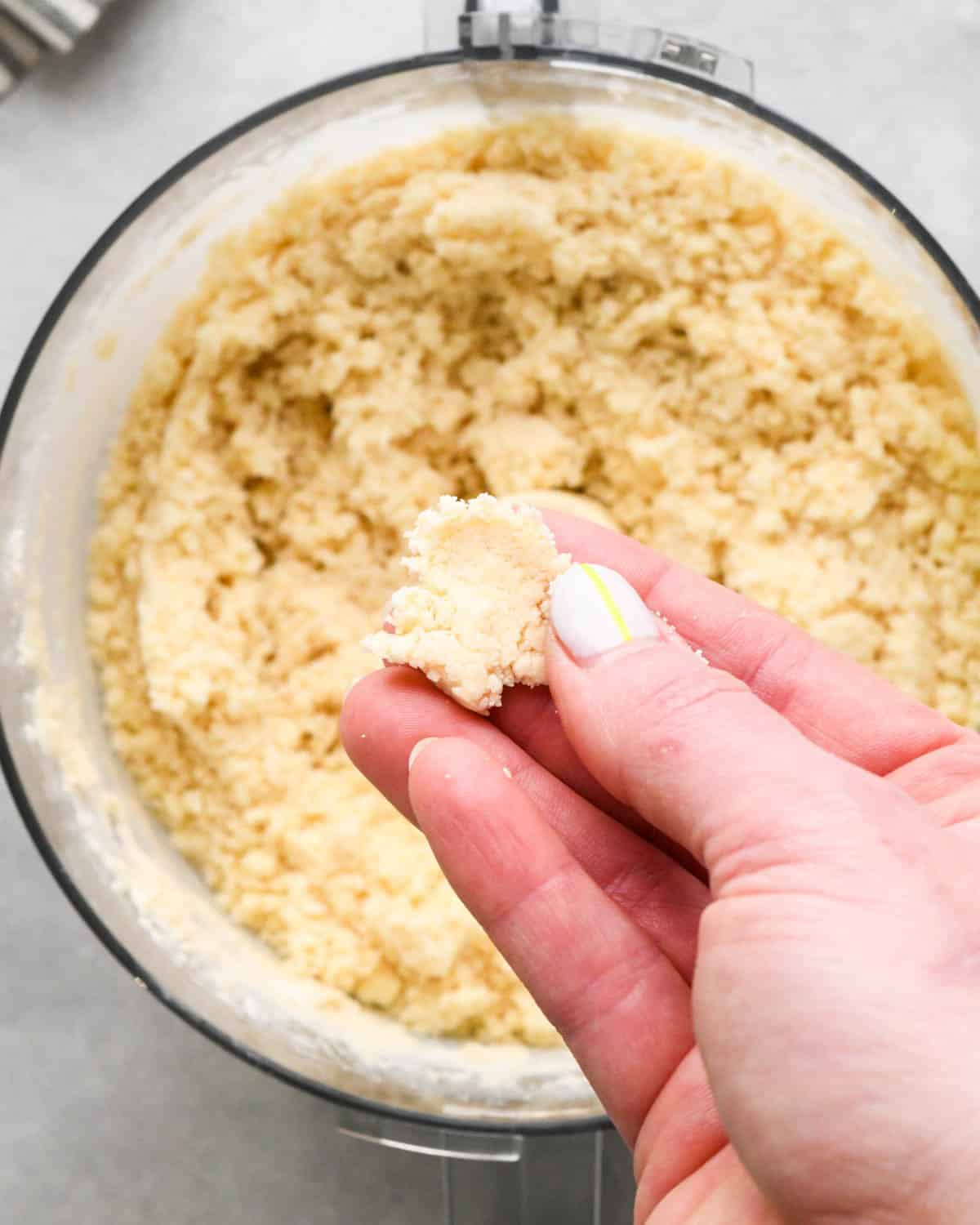 making crust for caramel apple pie - a hand holding a piece of pie crust mixture over the food processor that has been pinched together to show it's done