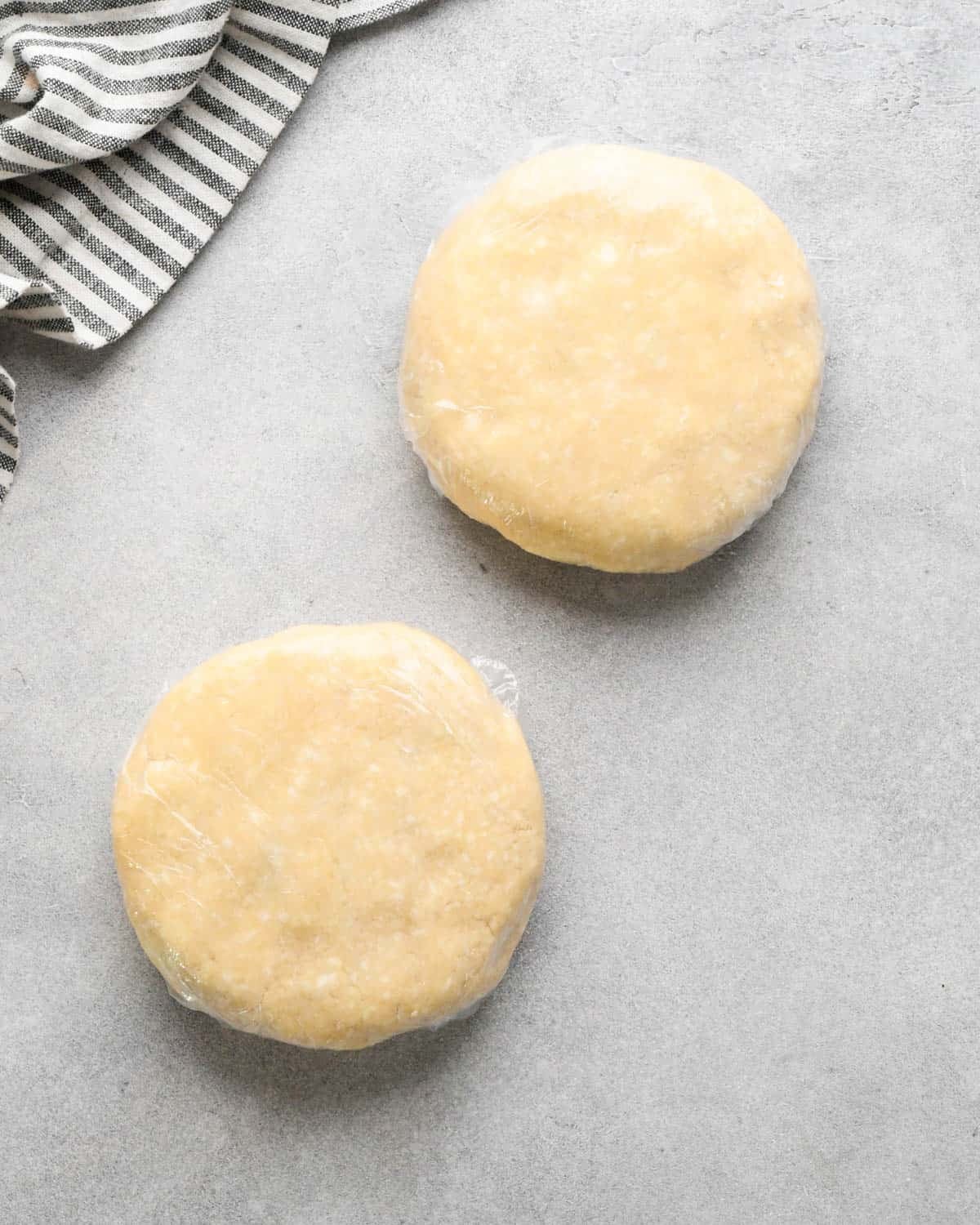 making crust for caramel apple pie - two discs of pie crust dough wrapped in plastic wrap on a gray surface, there's a striped towel int he top lefthand corner