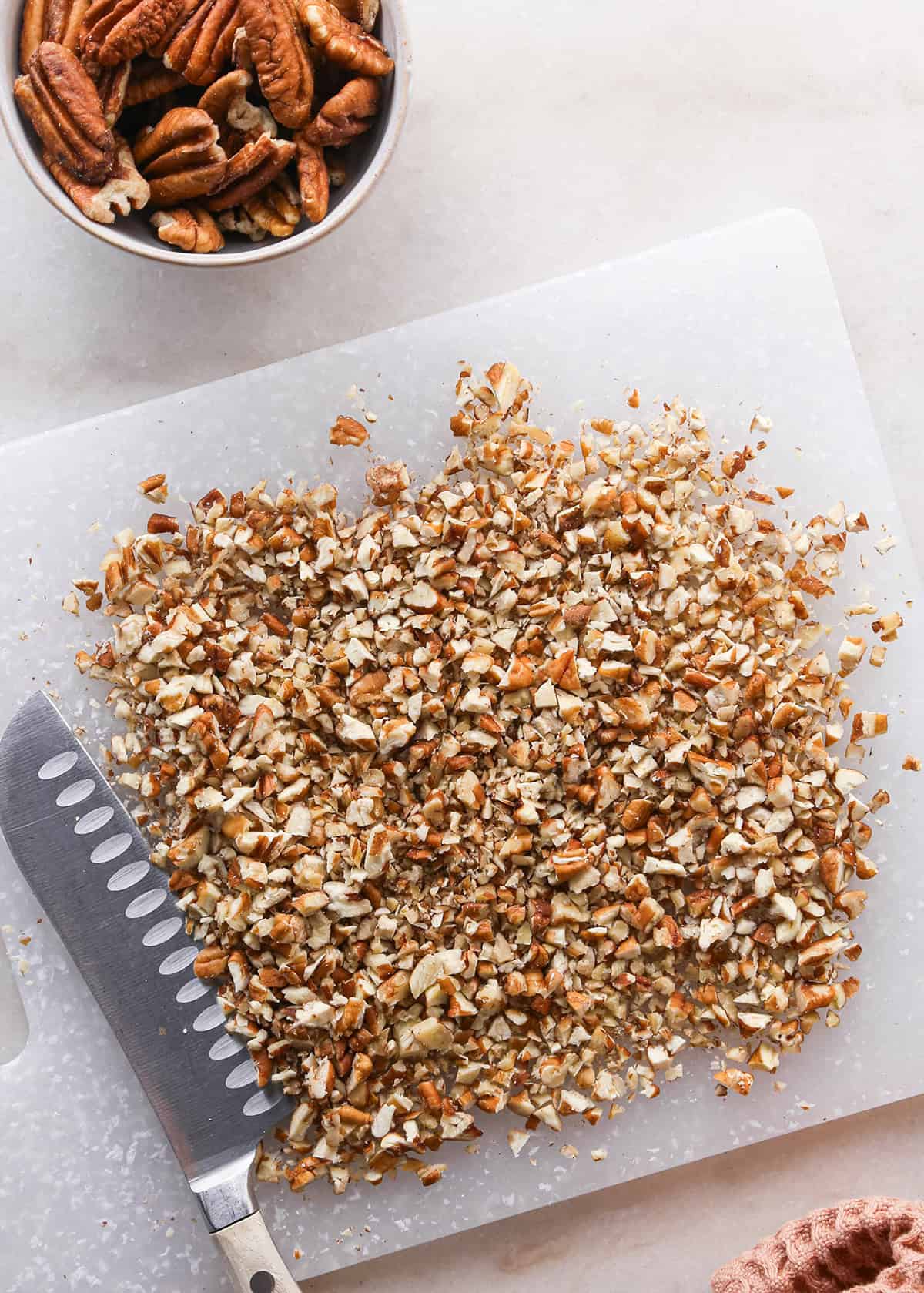 how to make pecan pie without corn syrup - chopped pecans on a cutting board with a knife, and a white bowl of pecan halves above it.