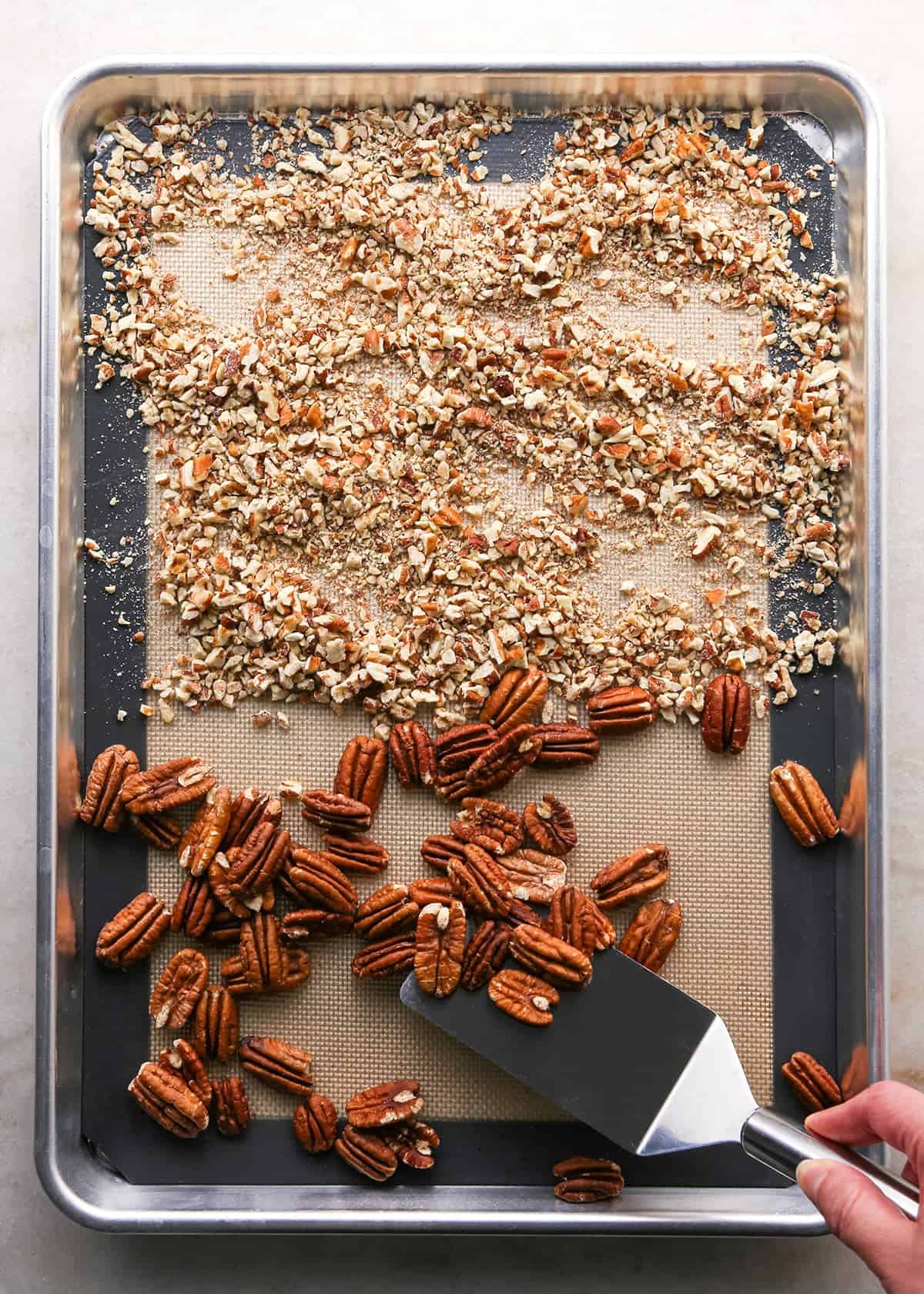 how to make pecan pie without corn syrup - chopped pecans and pecan halves on a large silver baking sheet lined with a silicone baking mat after being toasted in the oven, a hand holding a metal spatula is removing some pecan halves from the plate.