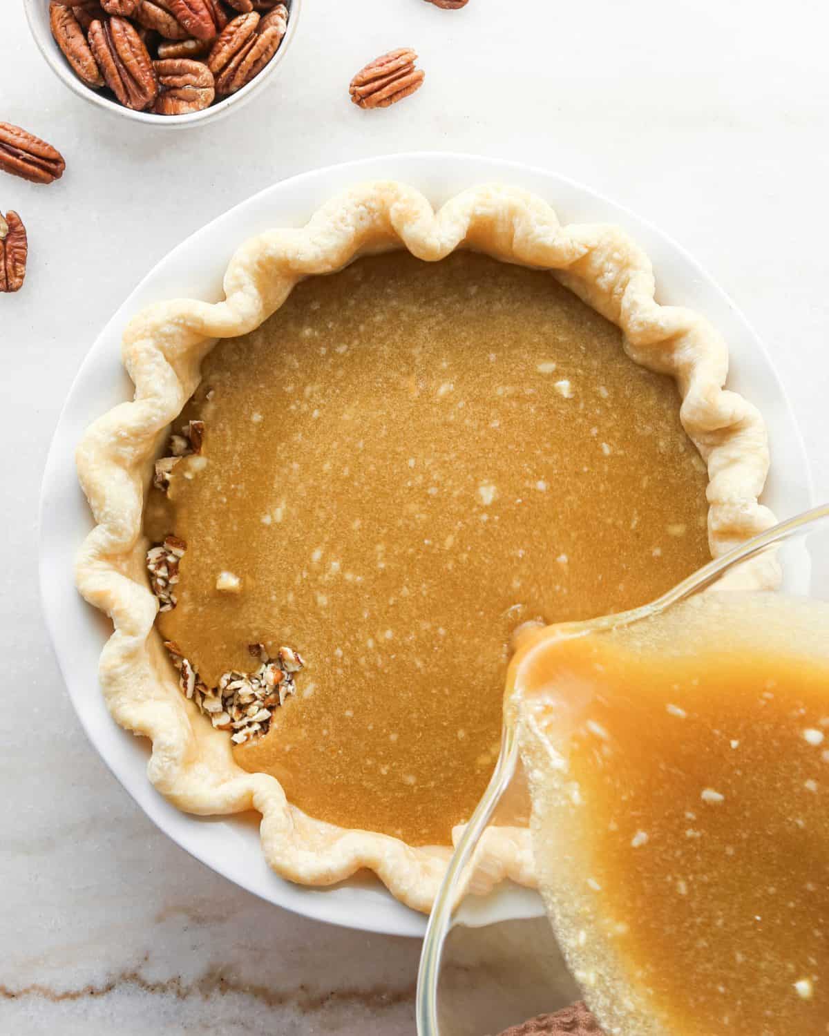 how to make pecan pie without corn syrup - pie filling being poured over the chopped pecans in the bottom of the partially baked pie crust in a white ceramic baking dish