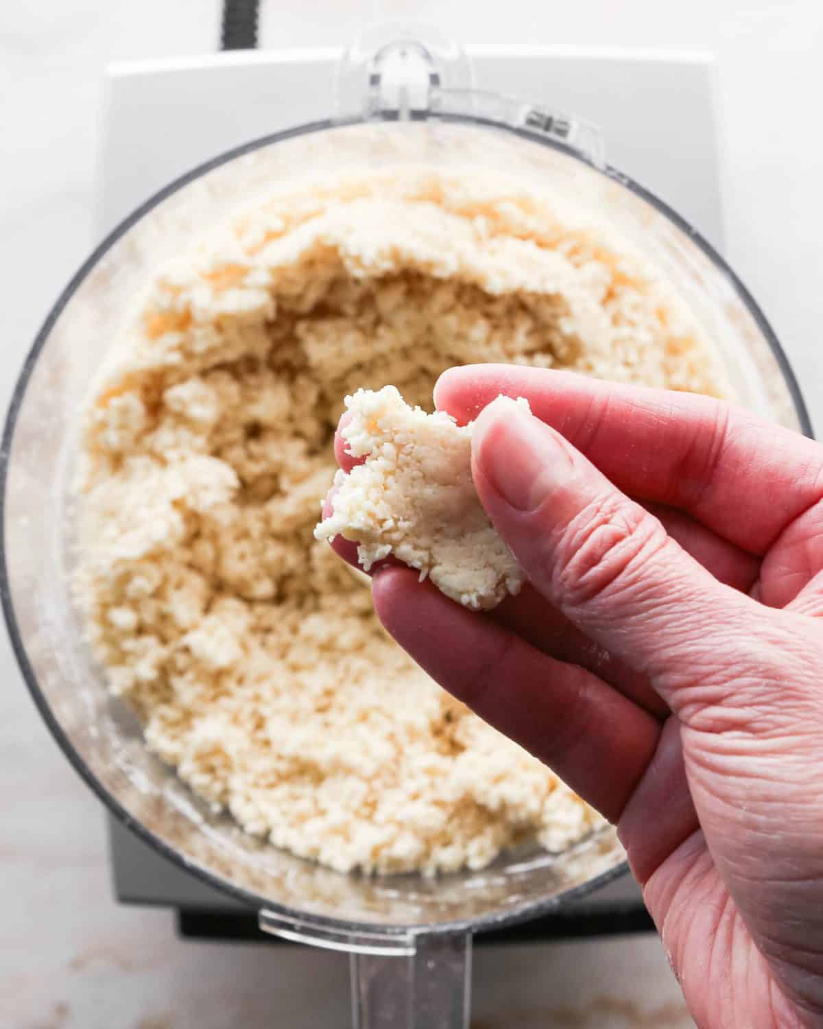 how to make pecan pie without corn syrup - a hand holding a pinched together piece of pie crust dough over the rest of the dough in a food processor fitted with the "S" blade.
