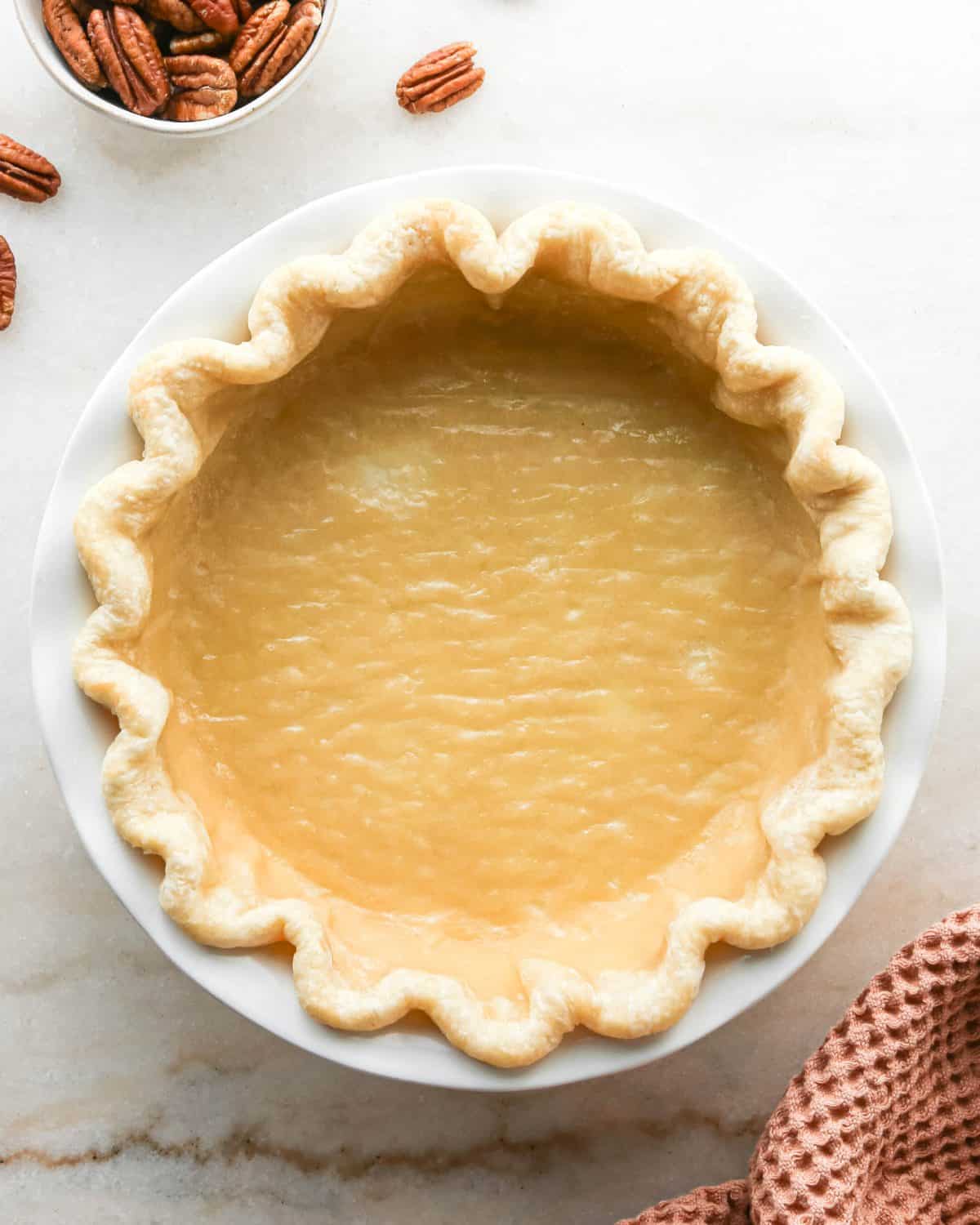 how to make pecan pie without corn syrup - pecan pie crust in the bottom of a white ceramic pie dish with a fluted edge after being partially baked