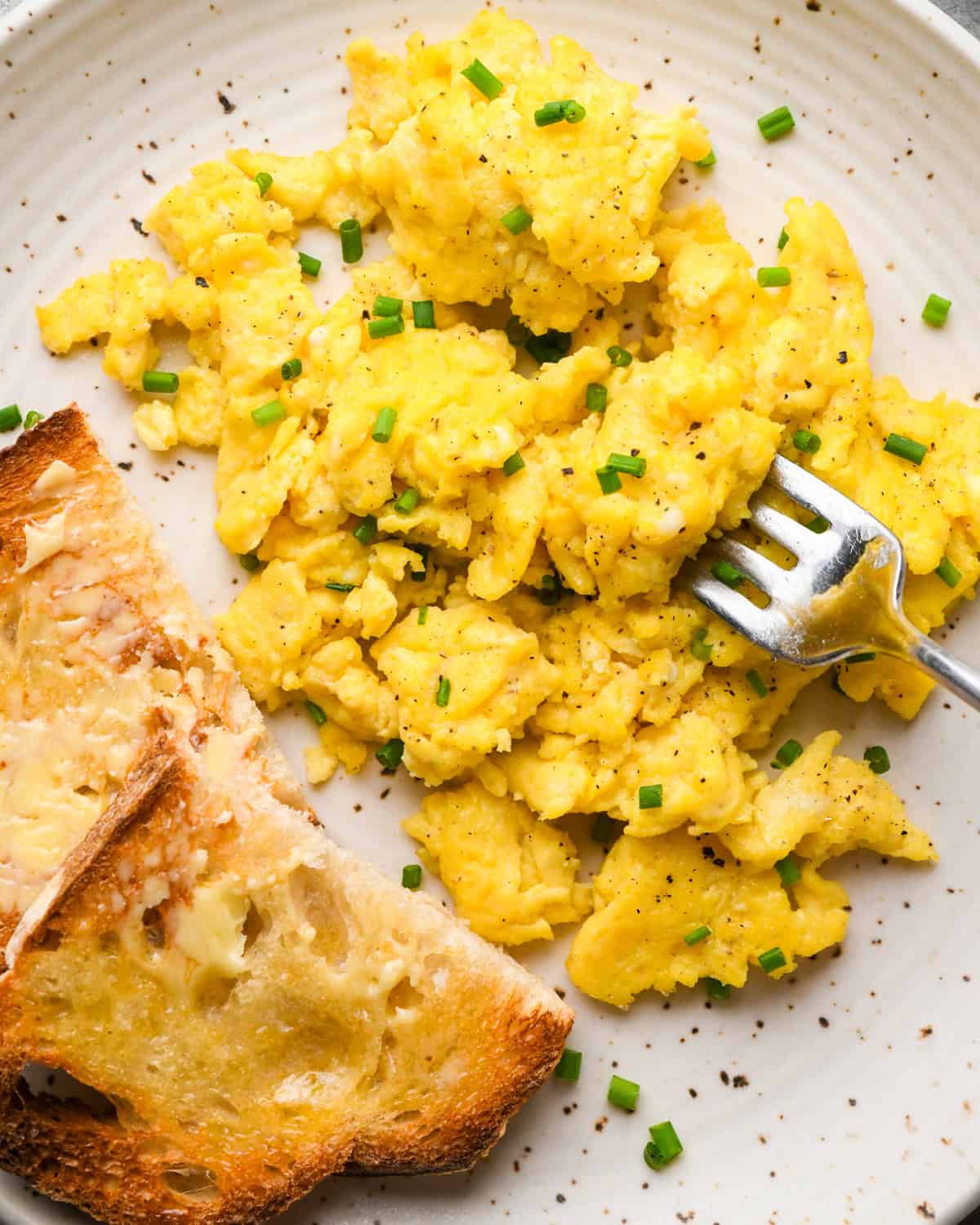 cottage cheese and scrambled eggs on a white plate garnished with chives and black pepper, with a piece of buttered toast on the side and a silver fork being stuck into the eggs