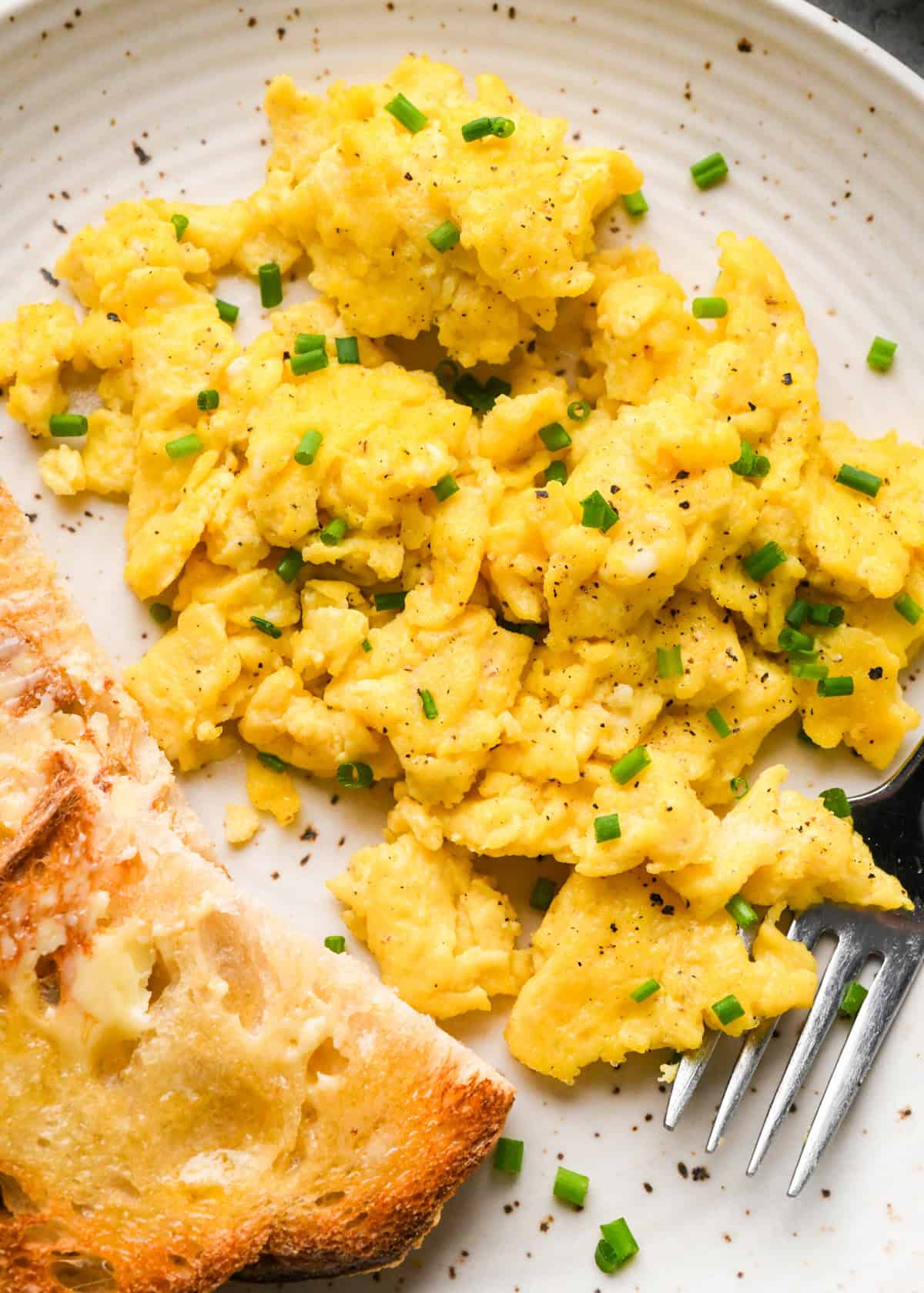 cottage cheese and scrambled eggs on a white plate garnished with chives, with a piece of buttered toast on the side and a silver fork on the plate. 