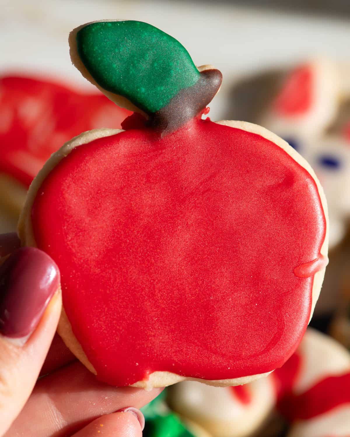 a hand holding an apple shaped sugar cookie decorated with sugar cookie icing that has hardened. The apple is red with a brown stem and green leaf. 