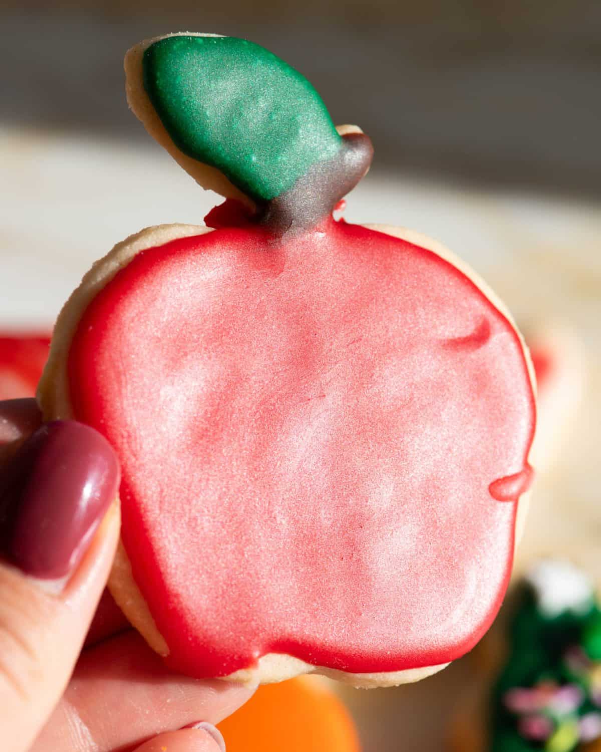 a hand holding an apple shaped sugar cookie decorated with sugar cookie icing that has hardened. The apple is red with a brown stem and green leaf. The cookie is tilted towards the sun so the shiny finish is visible. 