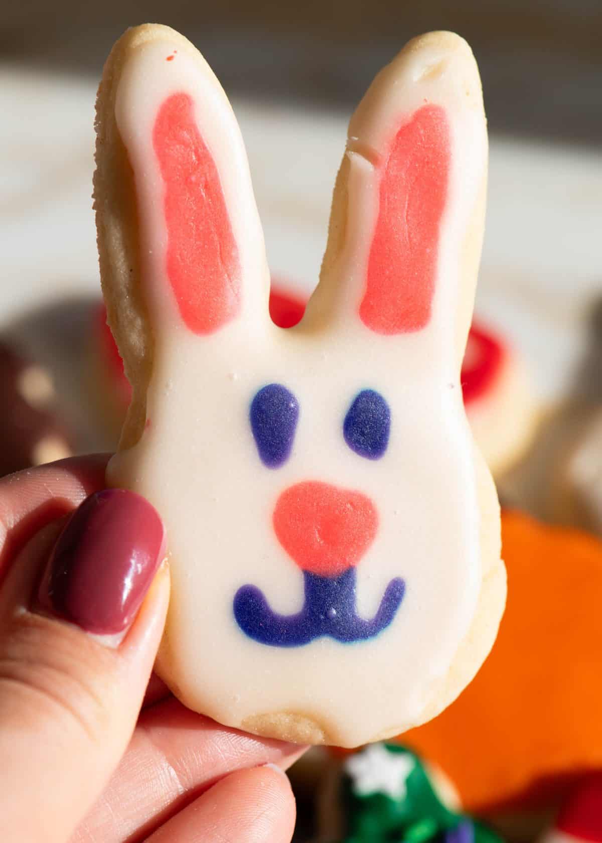 a hand holding a bunny shaped sugar cookie decorated with sugar cookie icing that has hardened. The bunny has pink ears and a pink nose, blue eyes and a blue mouth and it's face is white. 