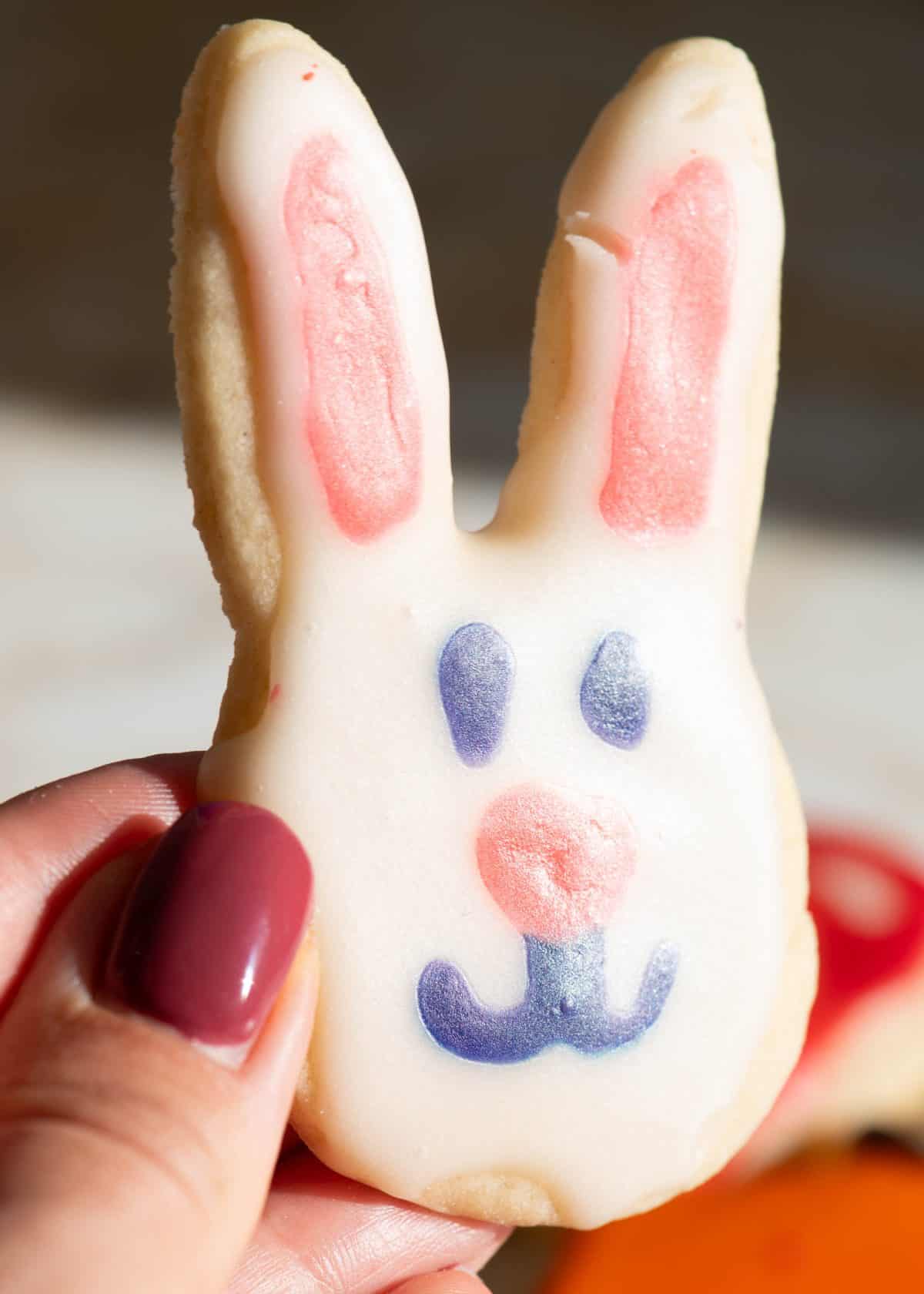 a hand holding a bunny shaped sugar cookie decorated with sugar cookie icing that has hardened. The bunny has pink ears and a pink nose, blue eyes and a blue mouth and it's face is white. The cookie is tilted towards the sun so the shiny finish is visible. 