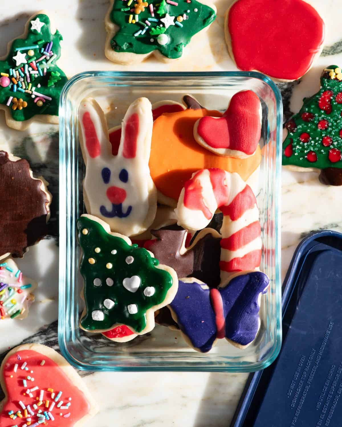 a variety of sugar cookies decorated with sugar cookie frosting that is shiny and has hardened in the shapes of bunnies, candy canes, butterflies, Christmas trees, pumpkins, turkeys and hearts. They are stacked in a glass container on a marble surface to be stored. 