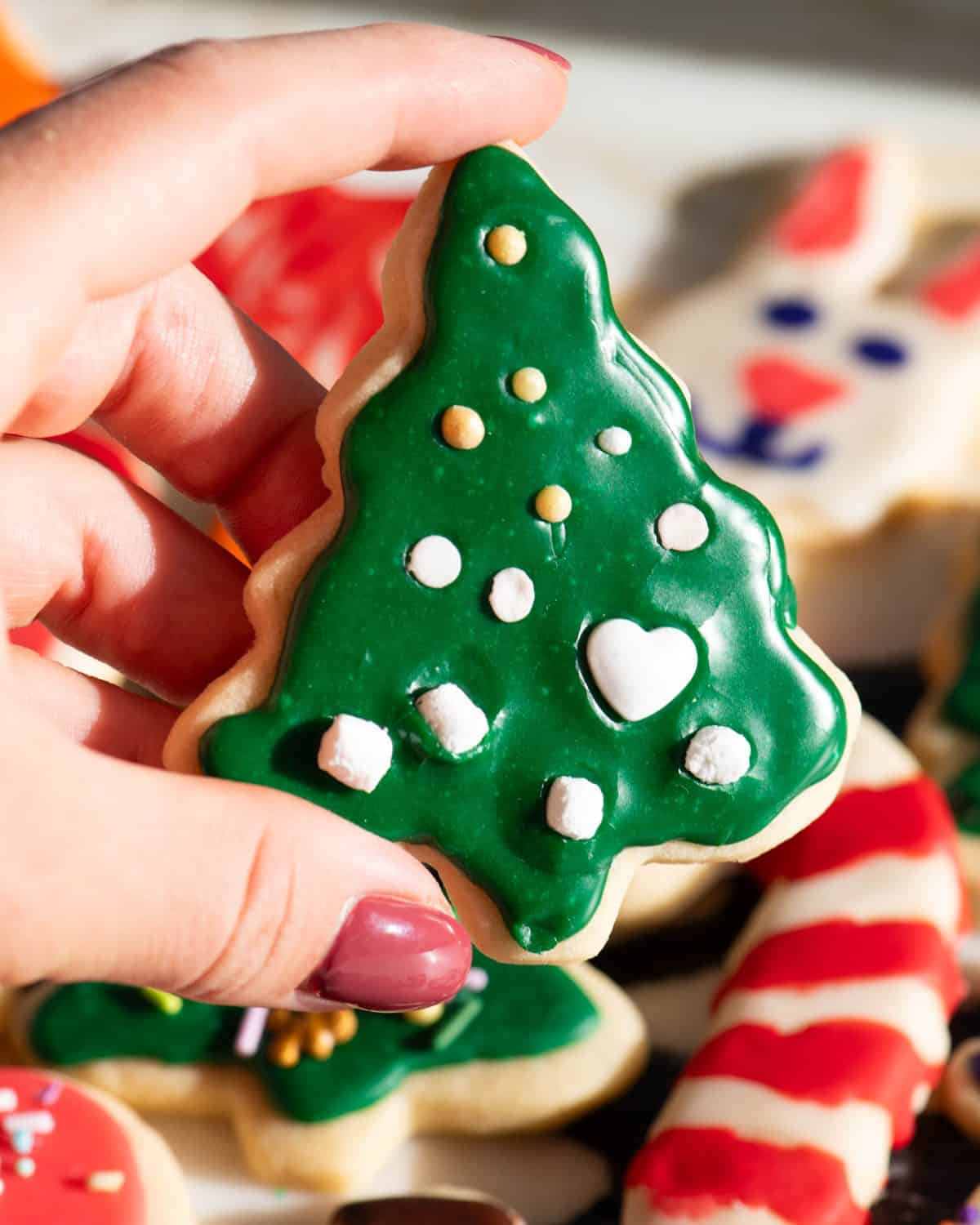 a sugar cookie Christmas tree with green sugar cookie icing on and white and gold sprinkles that look like ornaments. A hand is holding the cookie in the sunlight so you can see that it's shiny and there are more decorated sugar cookies in the background
