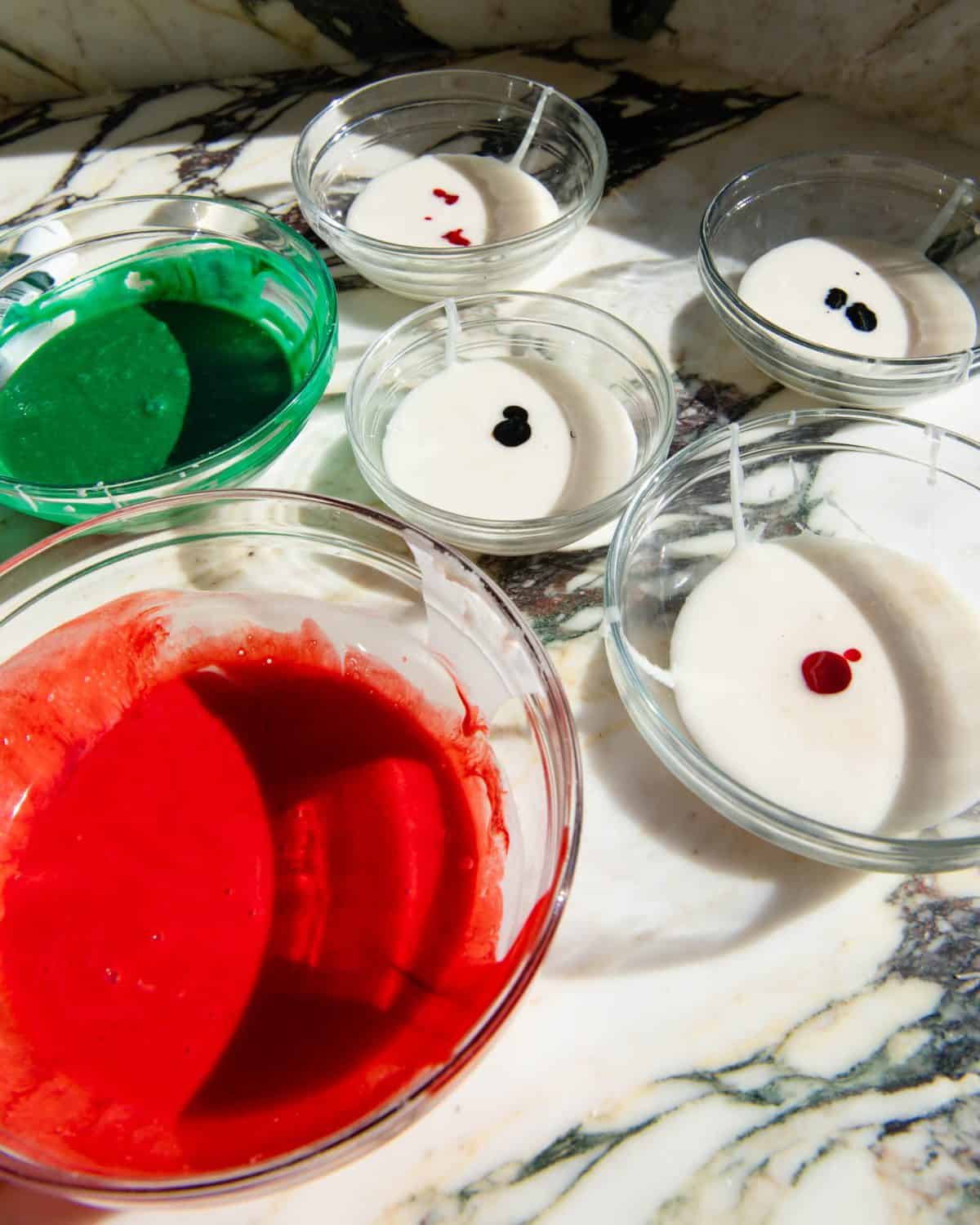 6 glass bowls of sugar cookie frosting on a marble surface. one has been dyed red, one has been dyed green, and the other 4 have drops of food coloring in them that hasn't been mixed. 