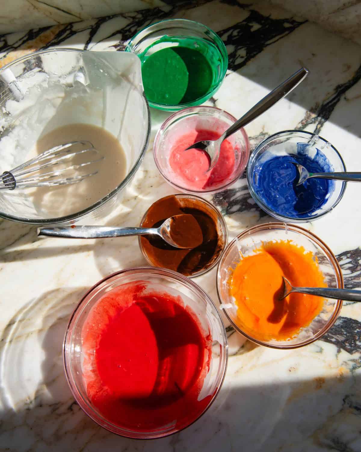 7 glass bowls of sugar cookie icing on a marble surface. 4 bowls have metal spoons in them. One has a metal whisk. They are dyed red, orange, brown, blue, pink, green and white 