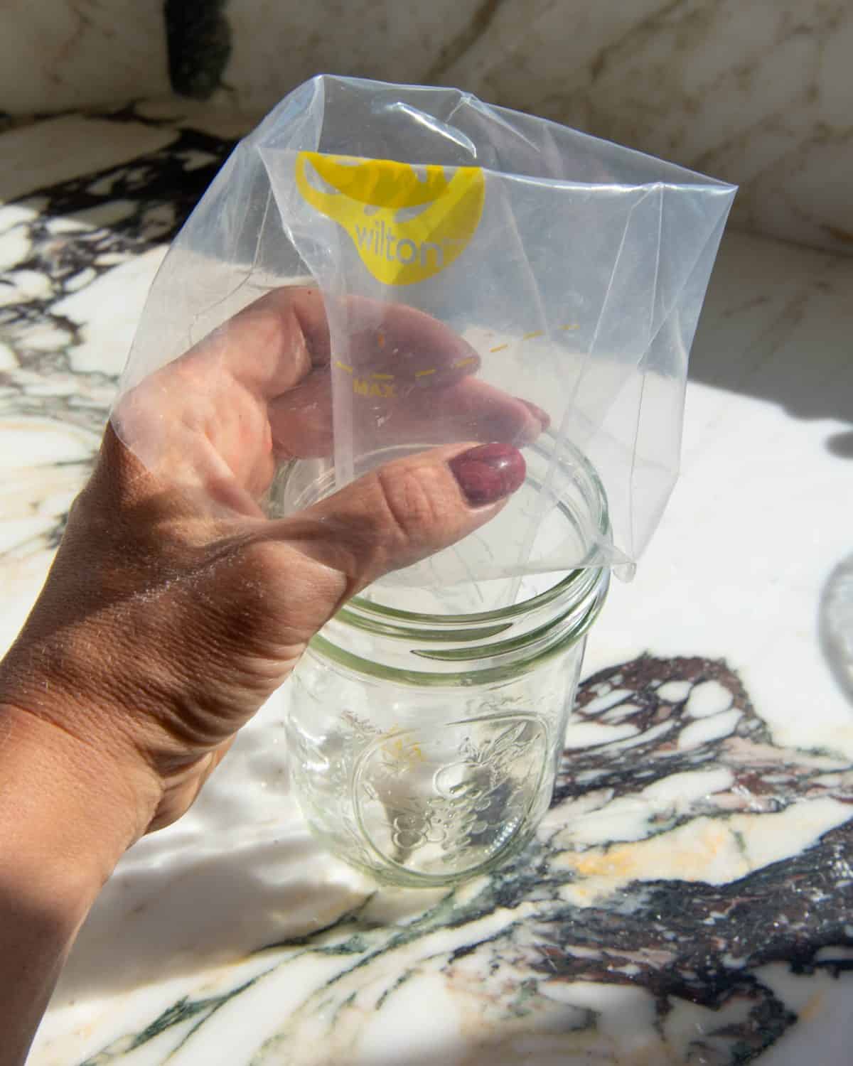 making sugar cookie icing - a hand holding a clear plastic piping bag in a glass jar on a marble surface
