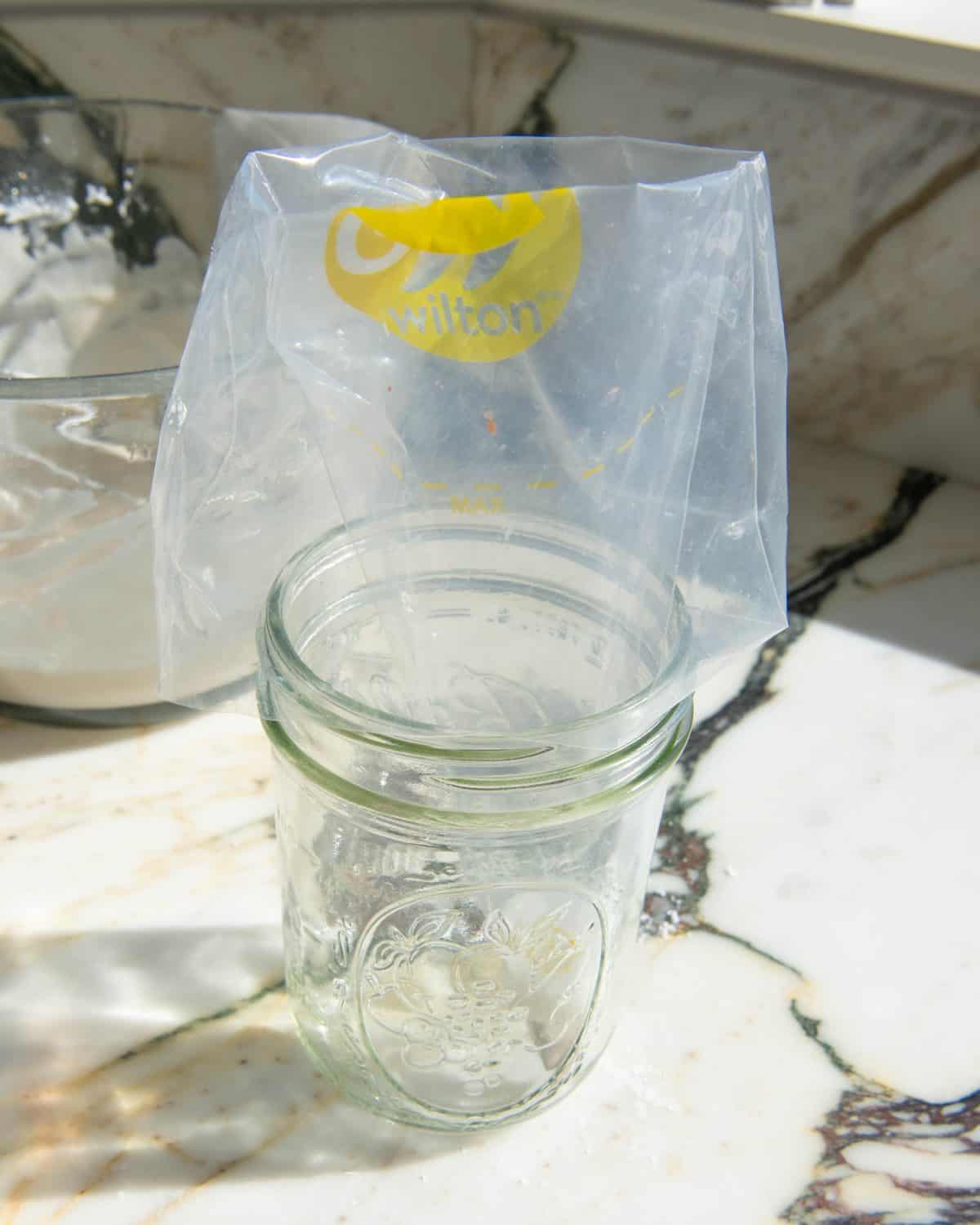 making sugar cookie icing - a clear plastic piping bag in a glass jar on a marble surface with a glass bowl of white sugar cookie frosting in the background