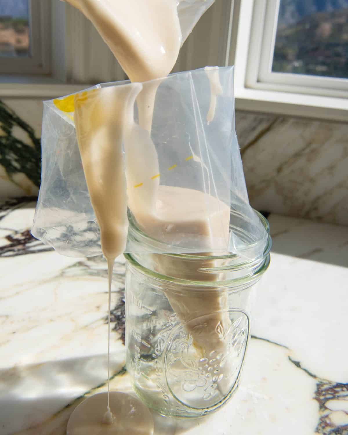 white sugar cookie icing being poured into a clear piping bag that's in a glass jar on a marble surface