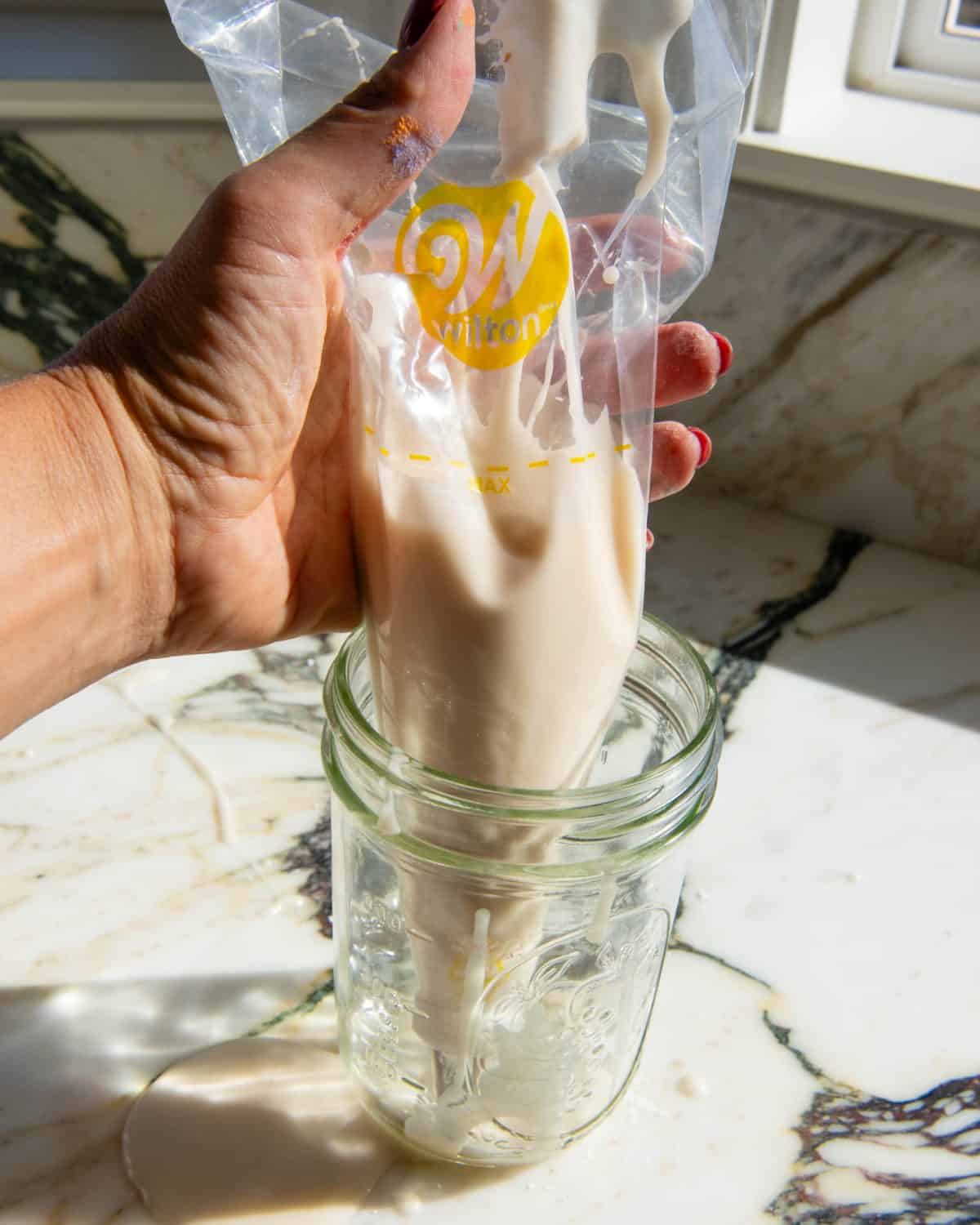 a hand holding a clear piping bag that's in a glass jar on a marble surface filled with white sugar cookie icing