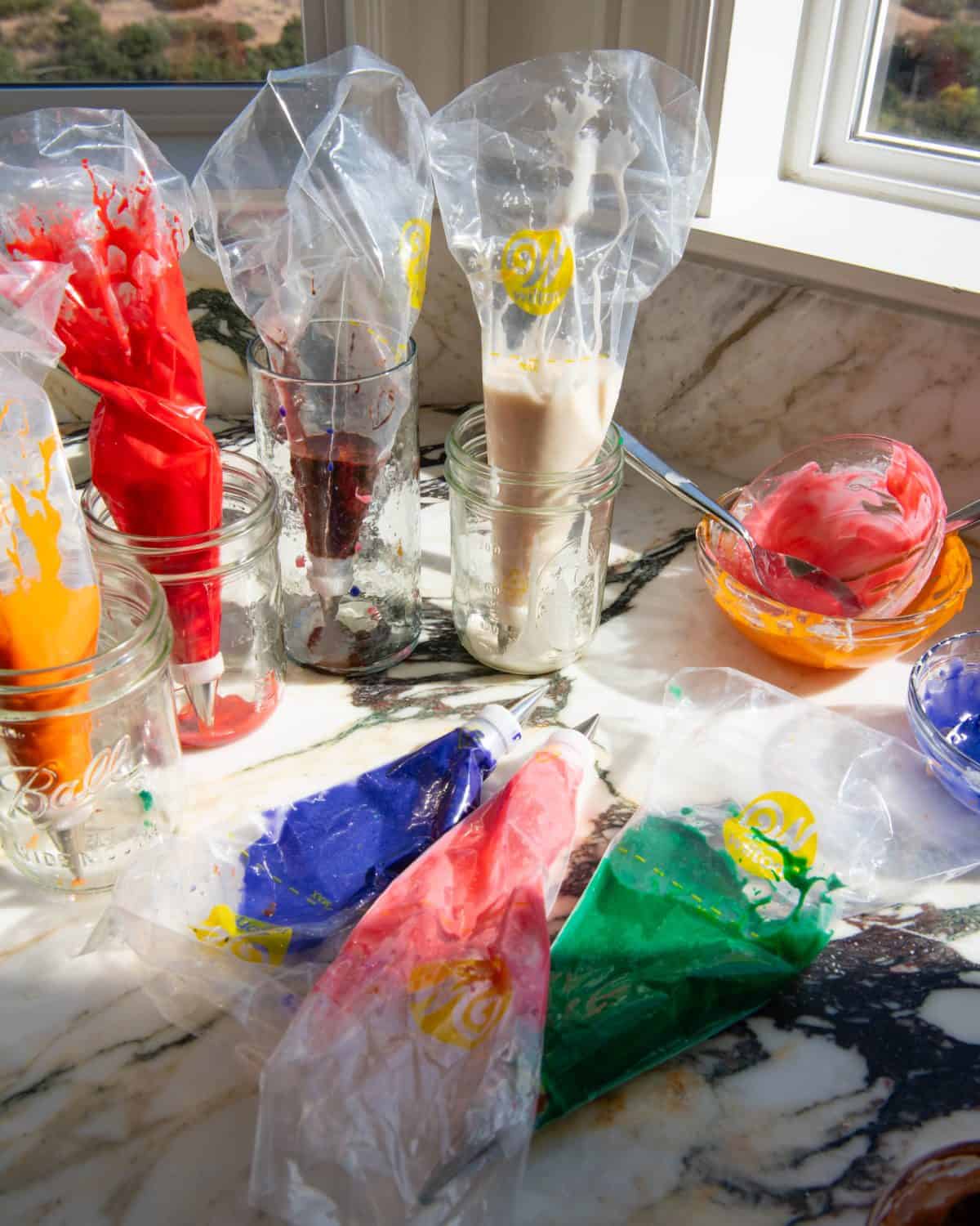 sugar cookie icing in 7 clear plastic piping bags, 3 bags are laying on a marble surface and they are blue, pink and green. 4 bags are standing up in glass jars and they are white, brown, red and orange. 