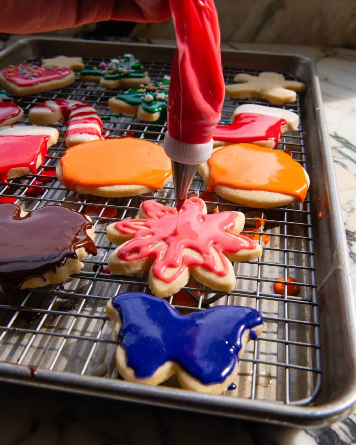 pink sugar cookie icing in a plastic piping bag with a metal tip decorating a flower sugar cookie that is on a wire rack in a baking sheet. there are more cookies decorated with sugar cookie frosting that are orange pumpkins, brown turkeys, candy canes and trees. 