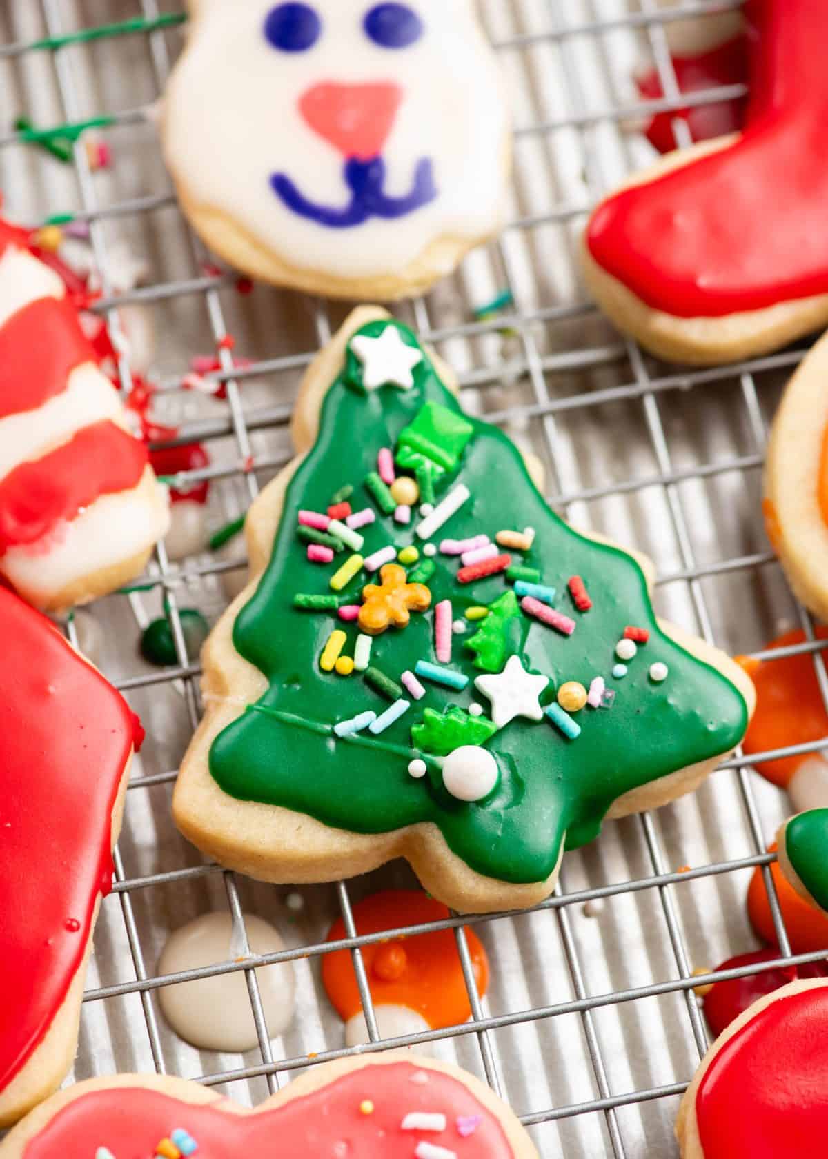 a Christmas tree sugar cookie that has been decorated with green sugar cookie icing and topped with sprinkles. The icing has hardened and is shiny. The cookie is on a wire rack surrounded by more cookies decorated with sugar cookie icing. 