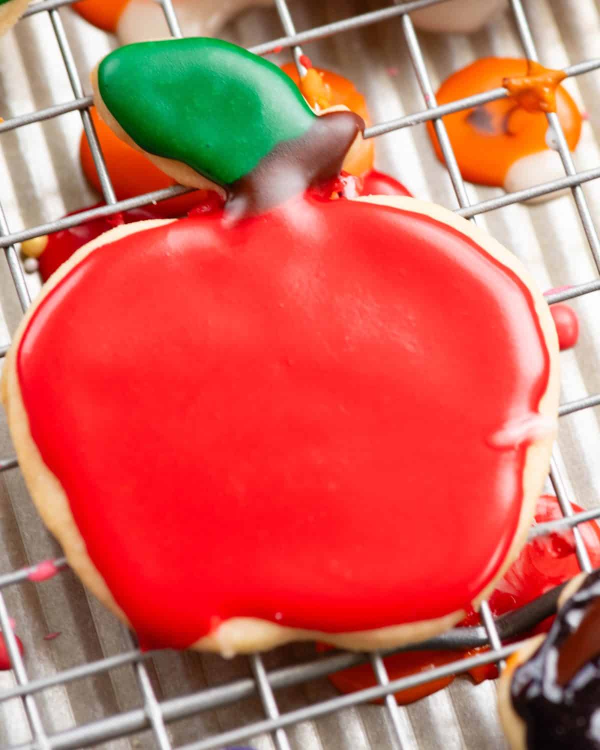 a sugar cookie in the shape of an apple decorated with sugar cookie icing with a brown stem and green leaf. The icing has hardened and is shiny. The cookie is on a metal wire cooling rack. 