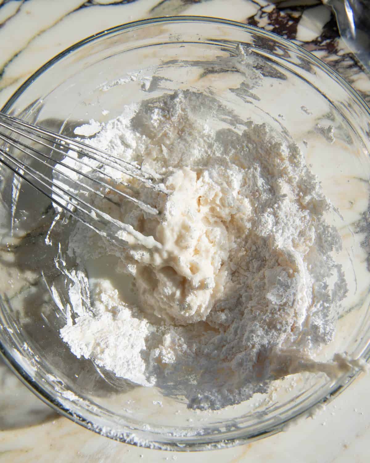 how to make sugar cookie icing - milk added to the ingredients in a glass bowl on a marble surface being whisked with a metal whisk