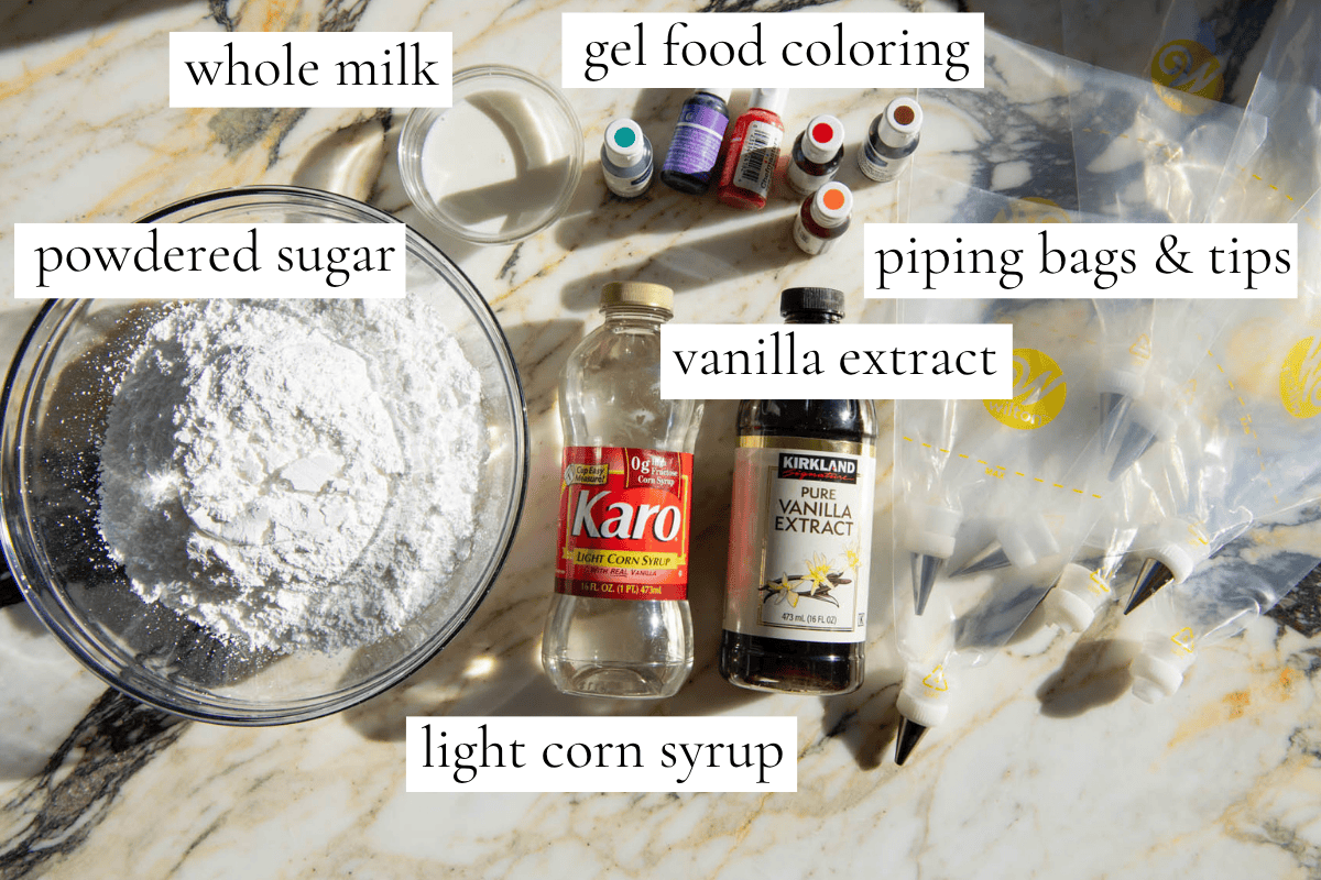 overhead photo of the ingredients in the sugar cookie icing recipe labeled, the ingredients are powdered sugar in a glass bowl, whole milk in a small glass bowl, gel food coloring, piping bags and tips, vanilla extract and corn syrup. 